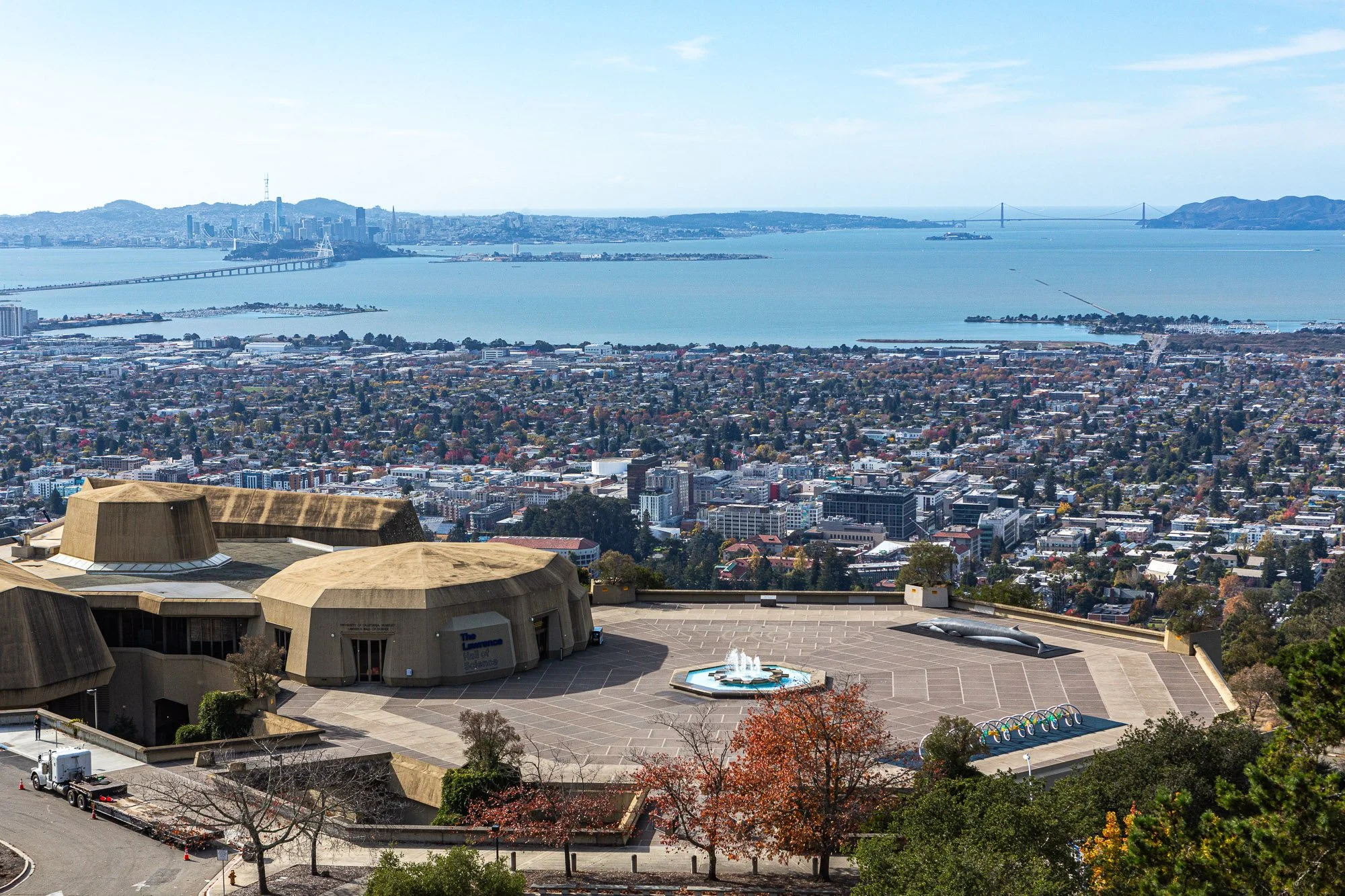 View of San Francisco skyline across the bay, with a large building with a rounded roof and a fountain in the foreground, and city streets and trees below.