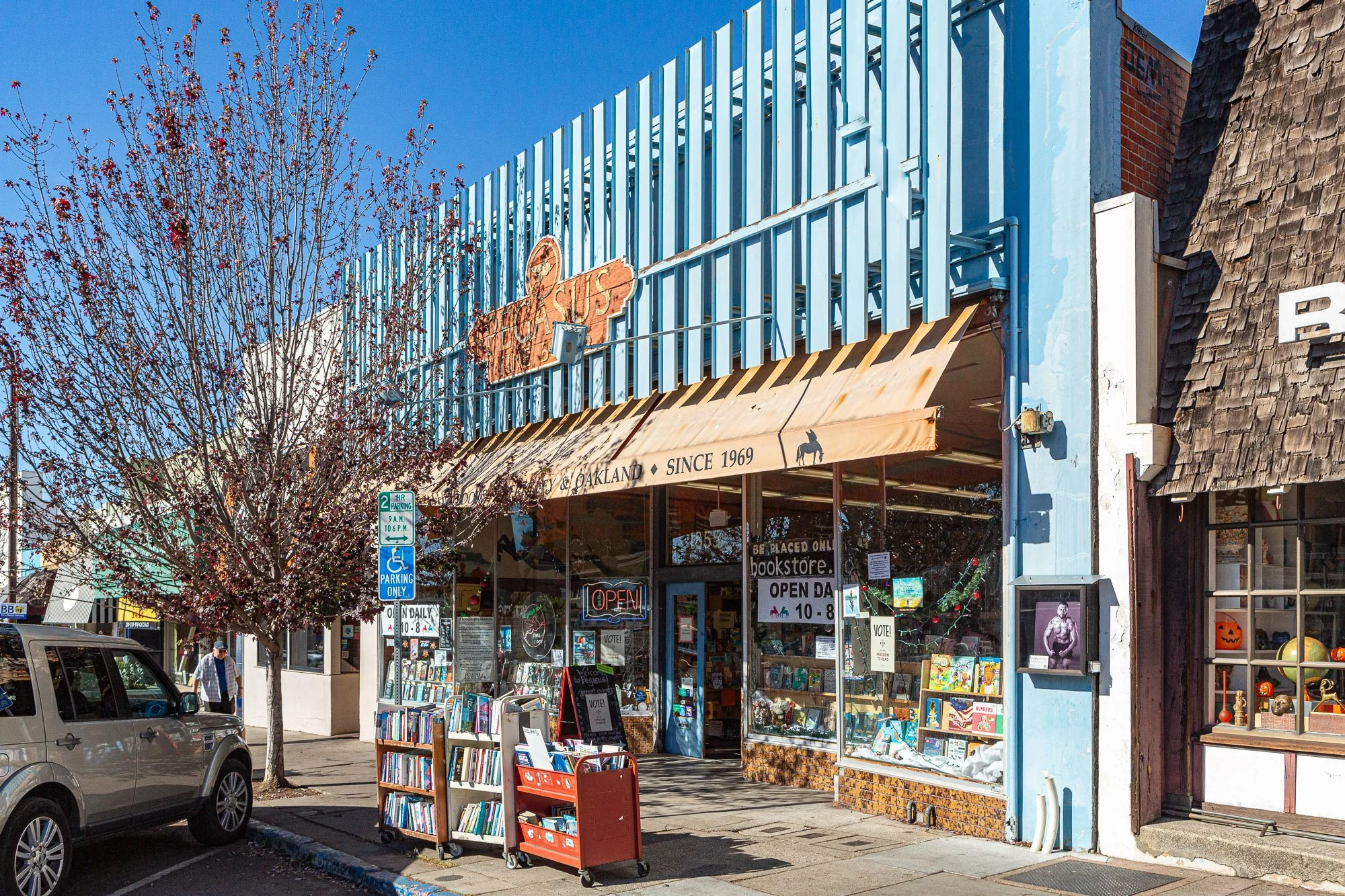 Bookstore located on a city street with a large front window and a beige awning that reads 'Books & Oakland Since 1969.' There are books displayed outside, a flowering tree, and a parked silver car. The store has signage indicating it is open daily f