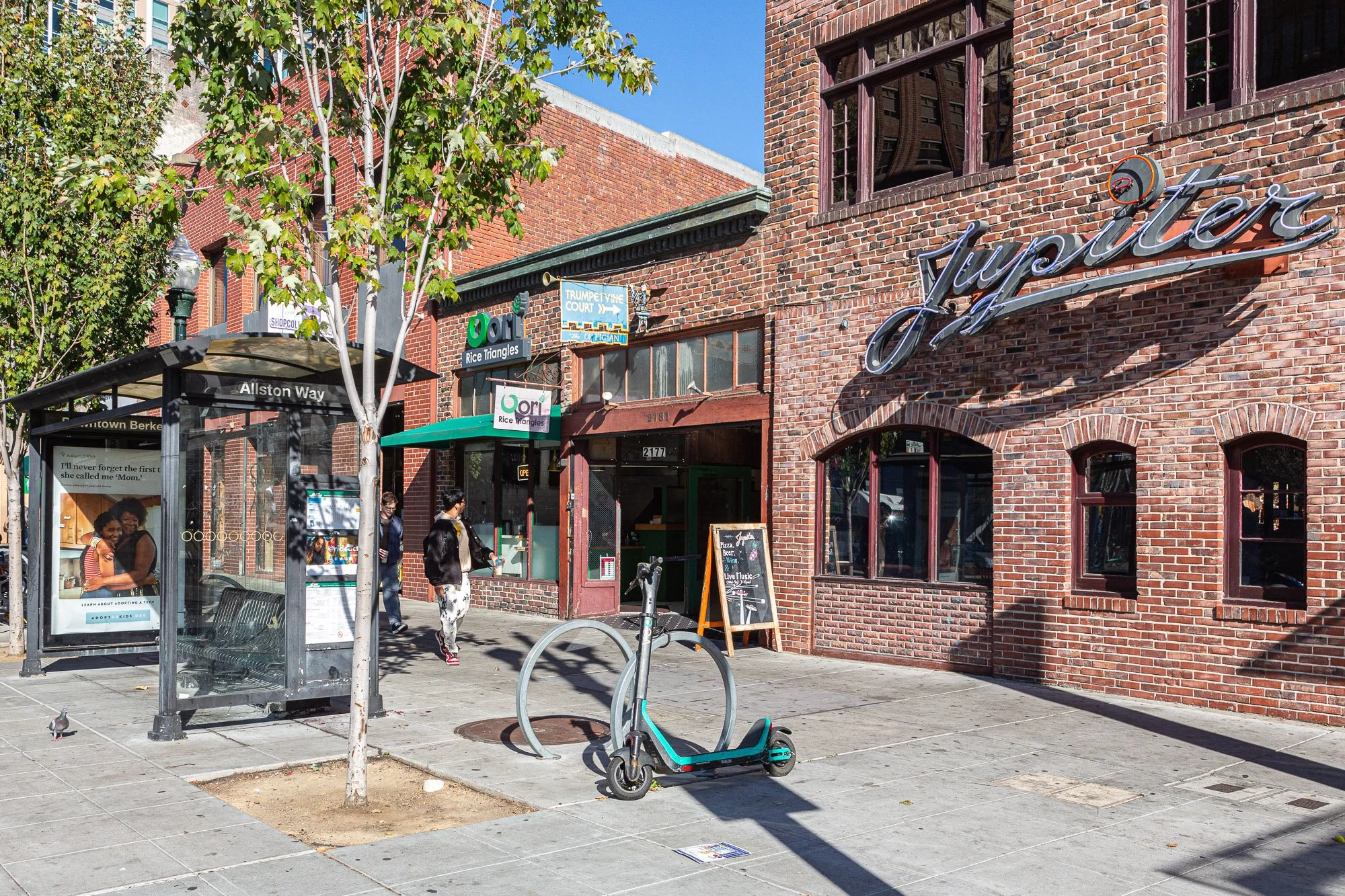 A city sidewalk scene with a brick building, a bus stop shelter, and a parked electric scooter in the foreground. People are walking near storefronts with signs, including 'Jazz' and 'Rice Triangles.' Trees and street furniture are also visible.