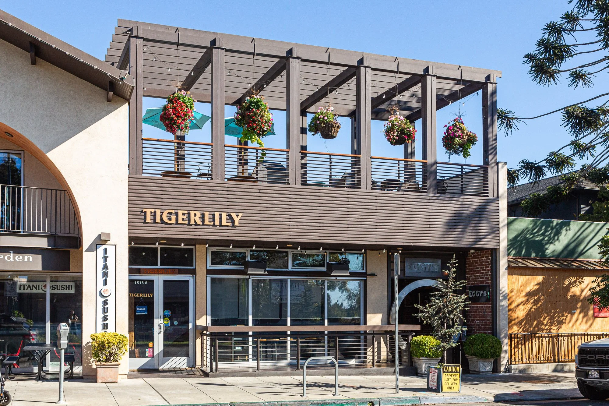 Two-story commercial building with a restaurant called Tigerlily on the ground floor and an outdoor seating area on the upper level with hanging flower baskets and umbrellas. The surrounding area has potted plants, signs, and parked cars.