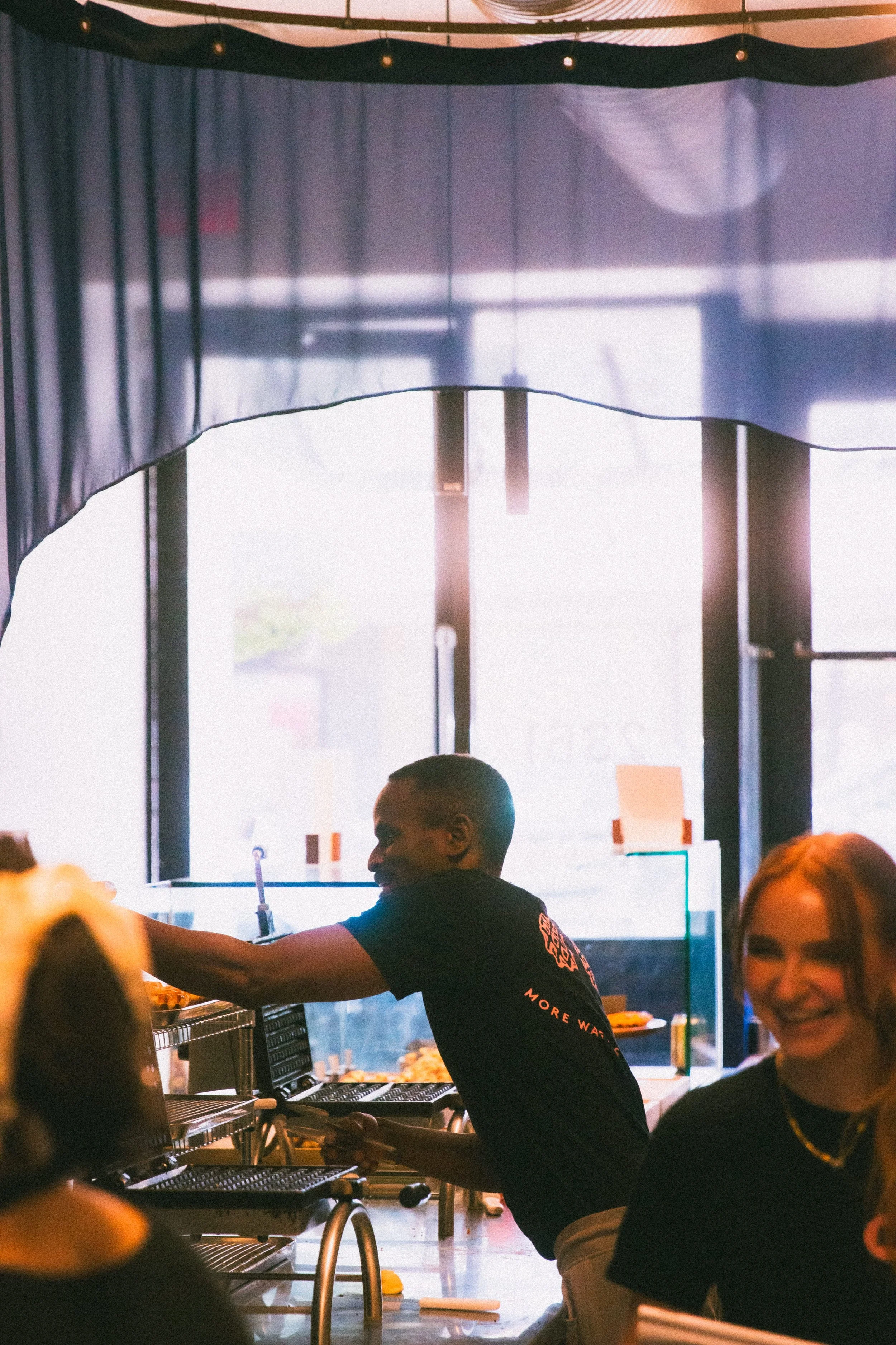 A person smiling and serving food at a food counter in a restaurant or cafe, with another person smiling in the foreground.