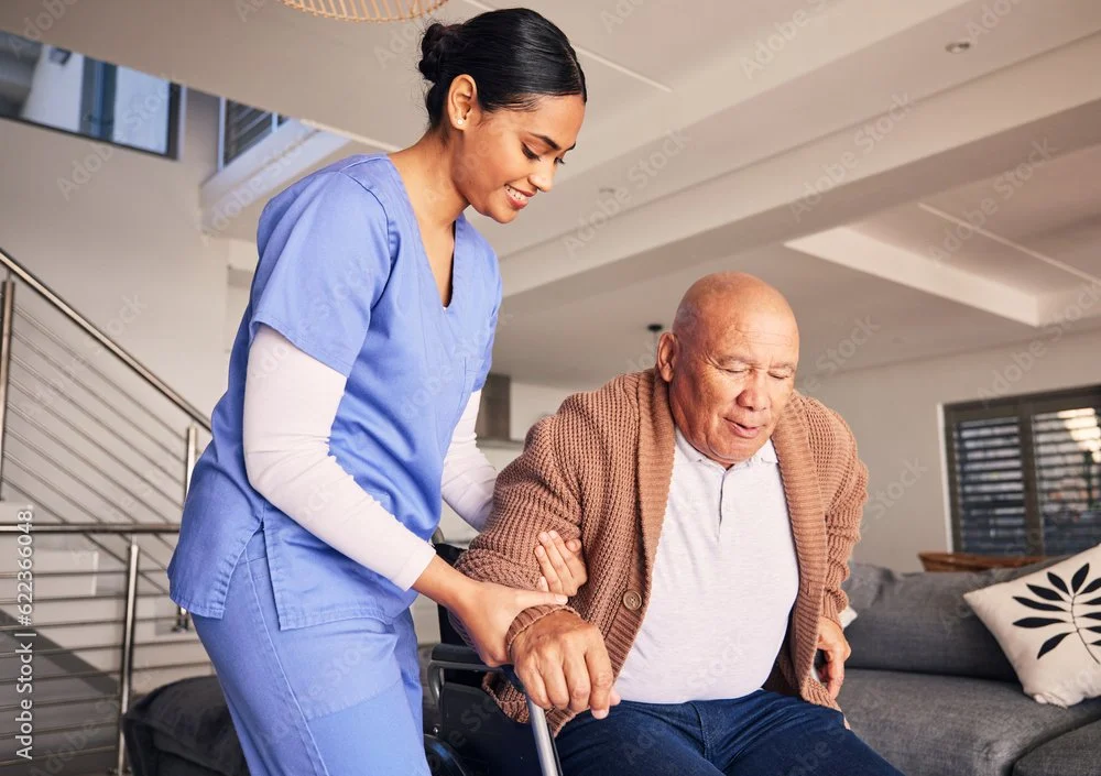A young female caregiver helping an elderly man in a wheelchair indoors, smiling and supporting him.