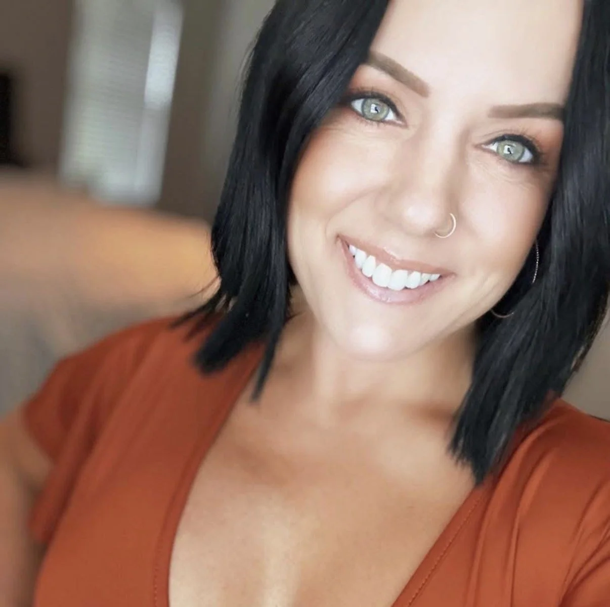 A woman with black hair, green eyes, and a nose piercing, smiling at the camera, wearing an orange top.