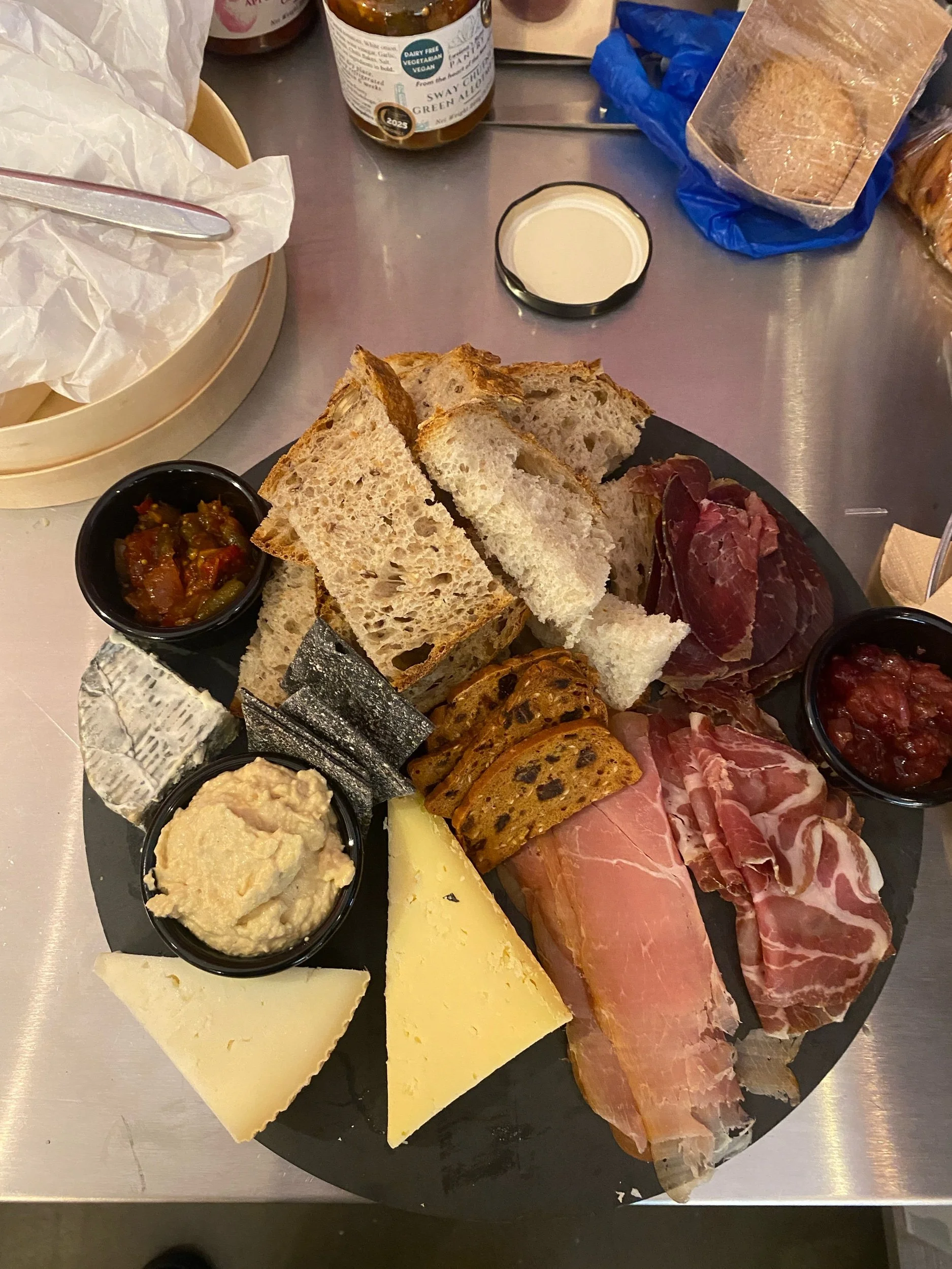 Charcuterie board with bread, cheeses, cured meats, spread, and condiments, set on a slate with additional chutneys, bread, and dips in the background.