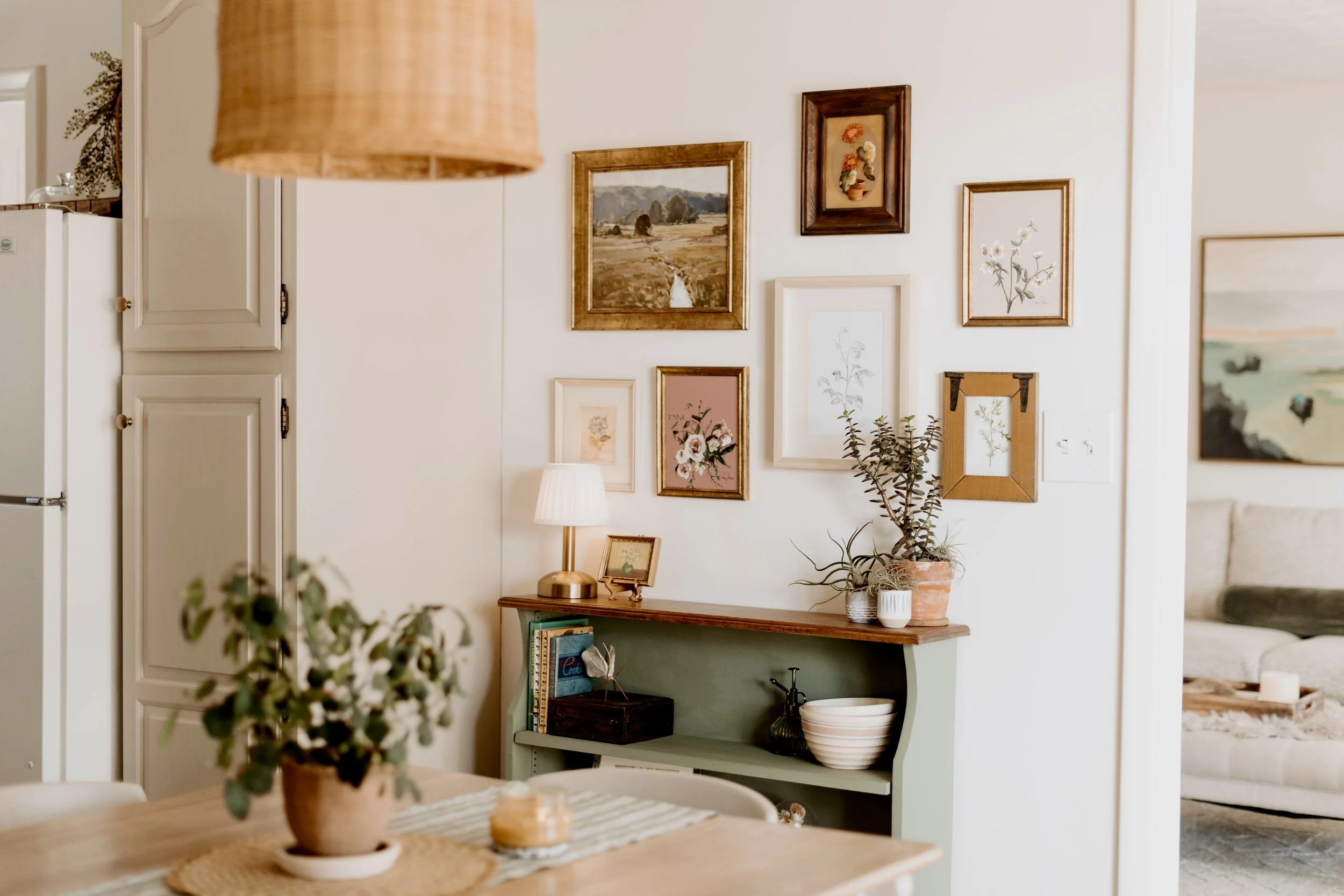 Interior of a cozy living space with a gallery wall of framed artwork, a small side table with a lamp, plants, and a bookshelf.