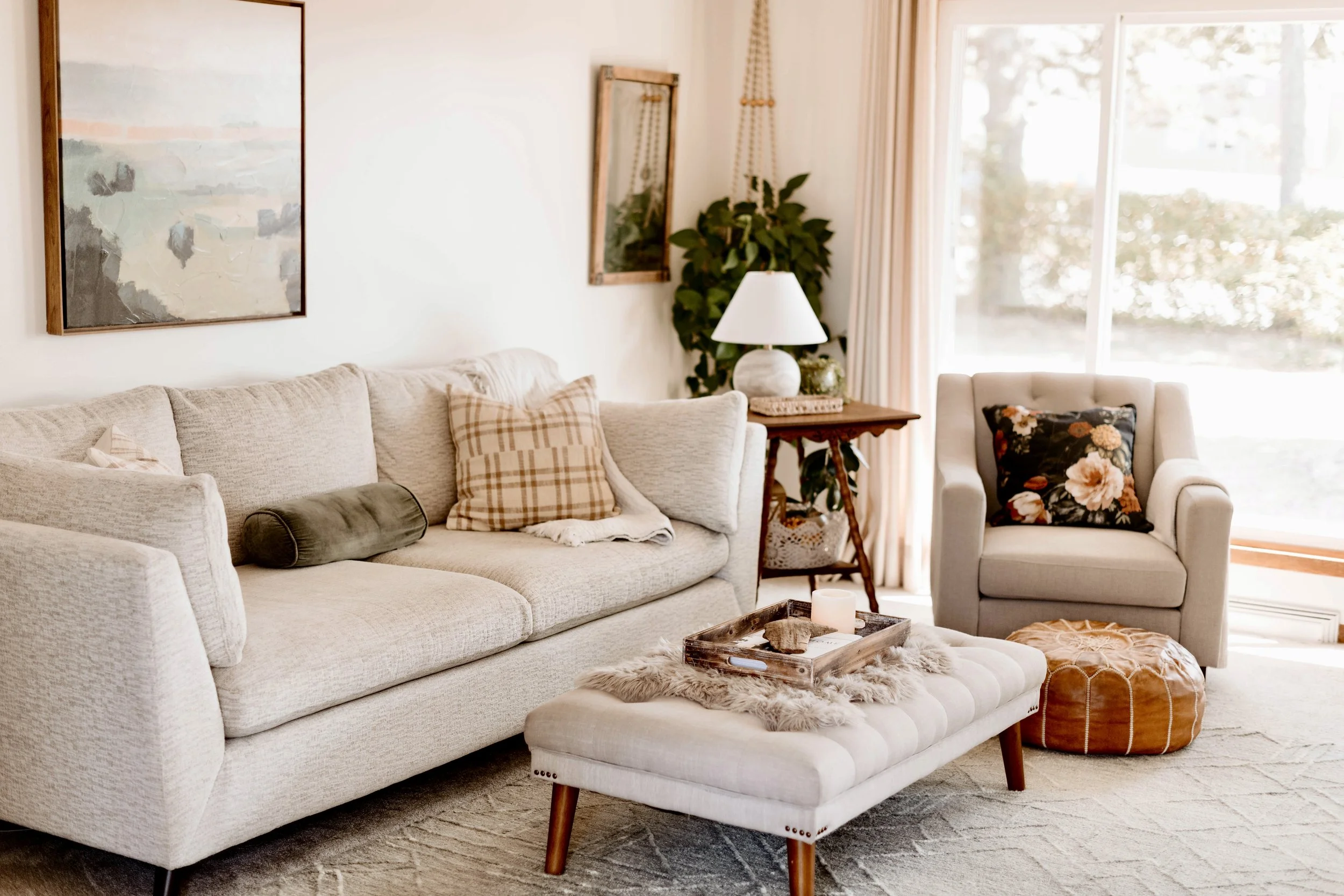 Living room with beige sofa, patterned throw pillows, white armchair with floral pillow, wooden side table with lamp, large window with curtains, and cozy decor.