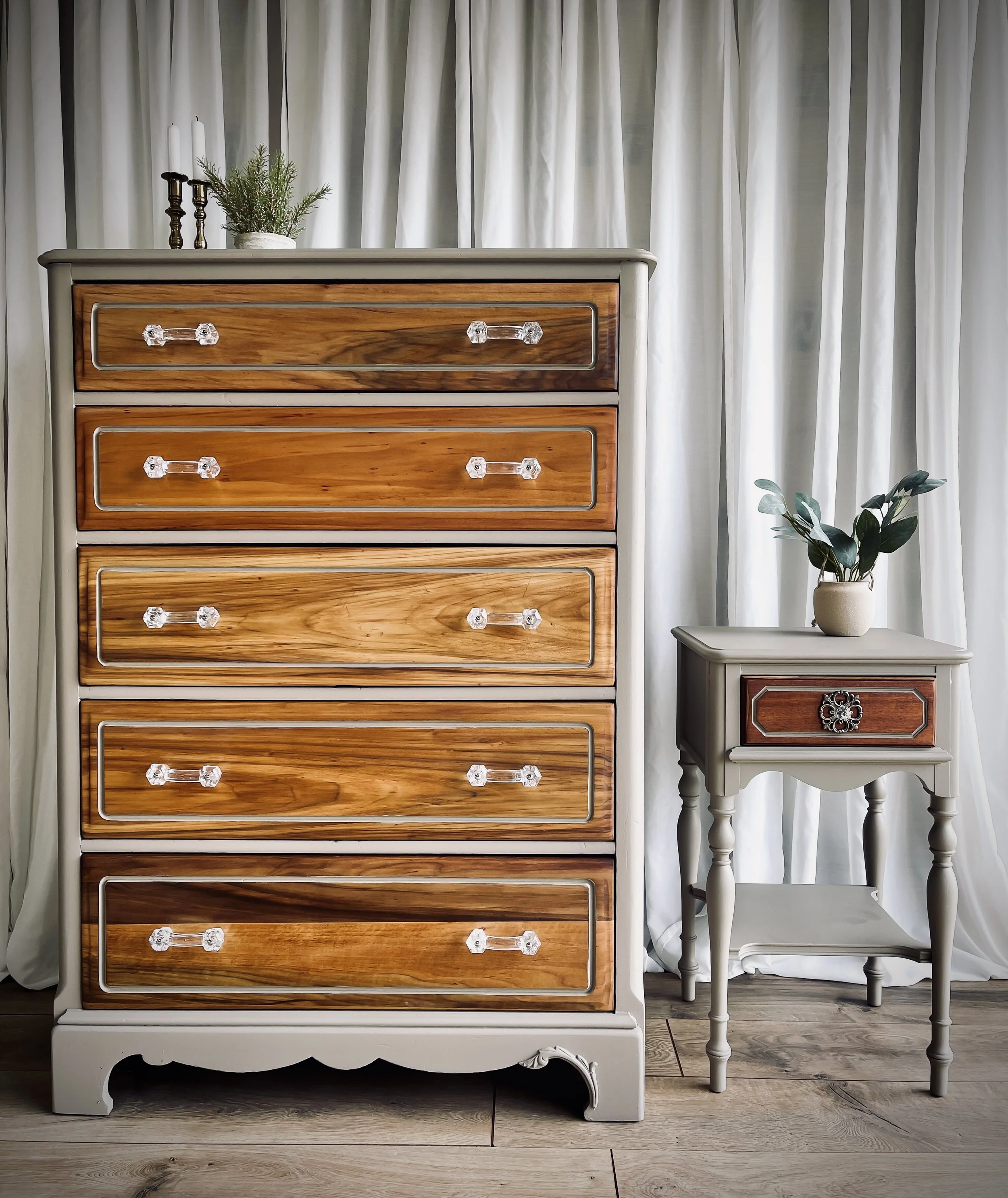 A wooden dresser with five drawers and clear glass knob handles next to a small gray side table with a drawer and decorative metal handle. The side table has a potted plant with broad green leaves. White curtains hang in the background.