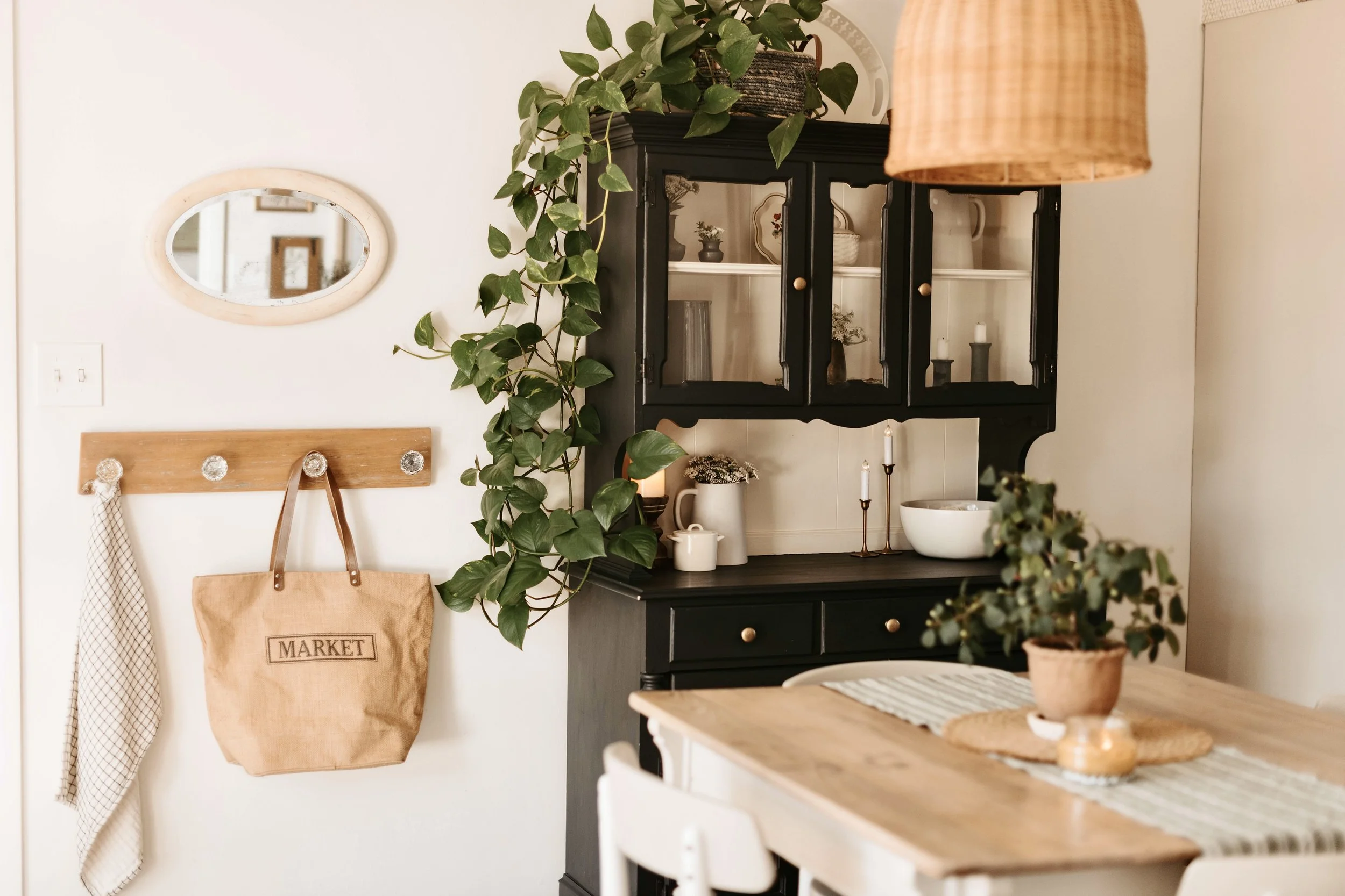 A cozy dining area with a black hutch decorated with plants, candles, and dishes, a wooden table with a striped table runner, and a small potted plant on top. A wall-mounted wooden coat rack with knobs holds a tan tote bag labeled 'MARKET' and a clot