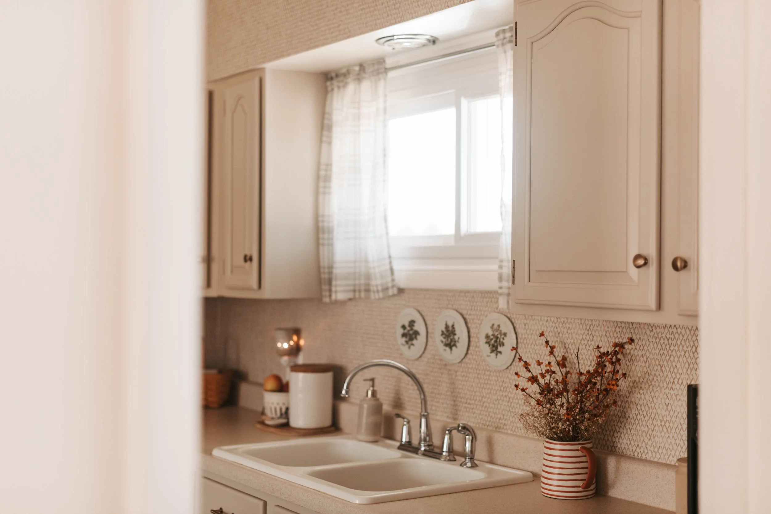Kitchen with beige cabinets, window with plaid curtains, white sink, red and white striped vase with dried flowers