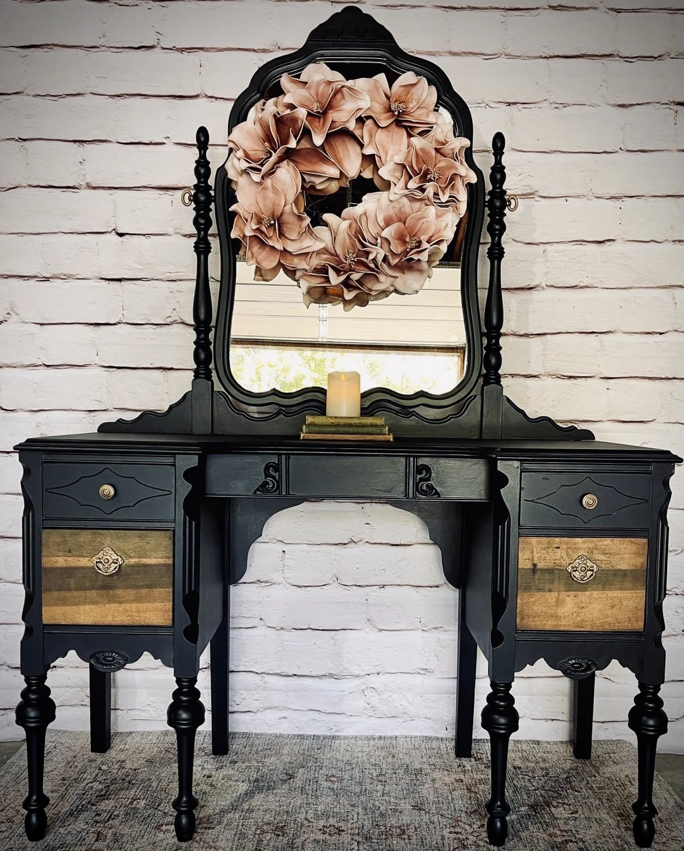 Black vintage vanity table with a large mirror, decorated with a pink floral wreath hanging on the mirror, a lit candle, and a small stack of books, set against a white brick wall.