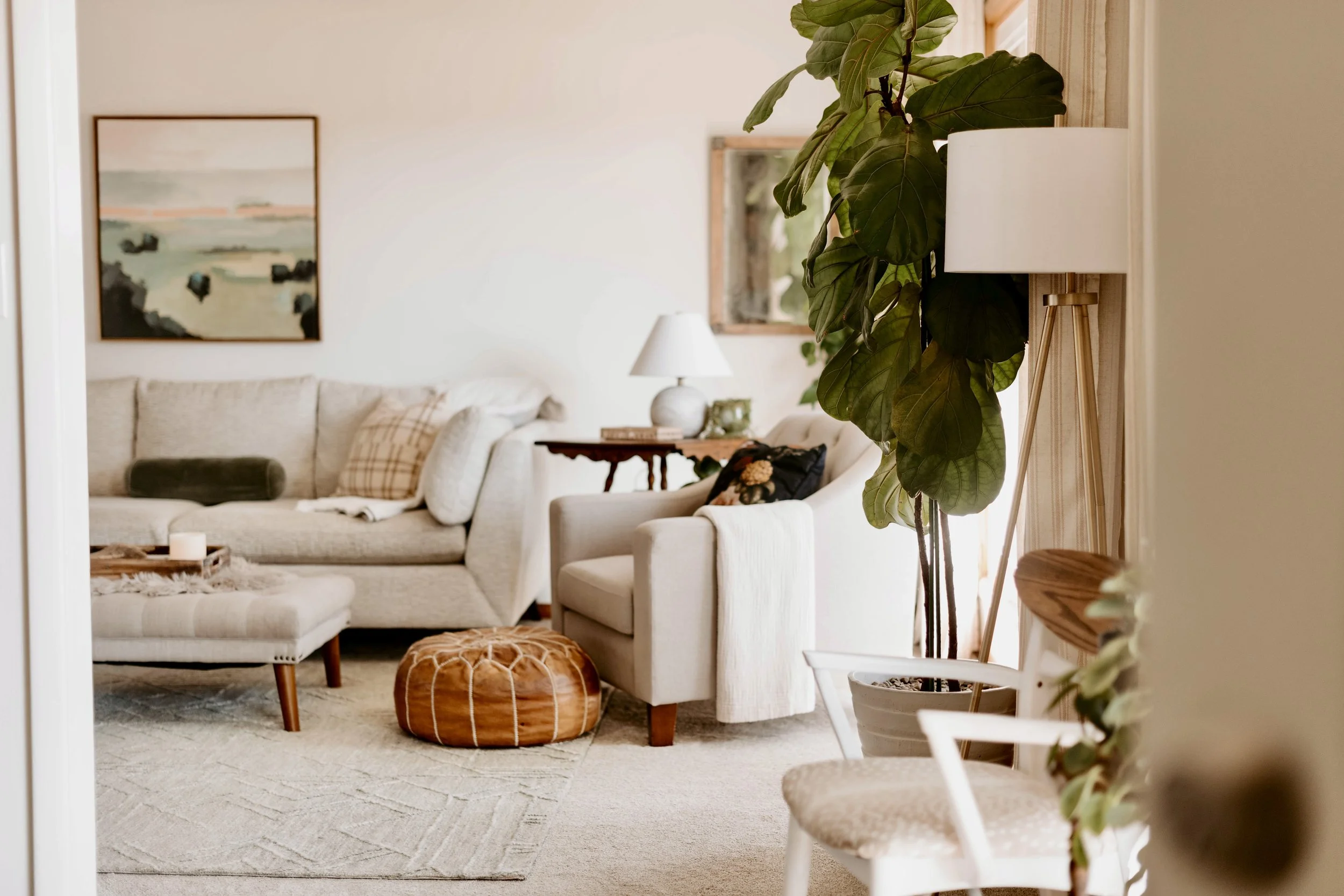 Living room with beige sofa, white armchair, wooden side tables, table lamp, indoor plant, and wall art.