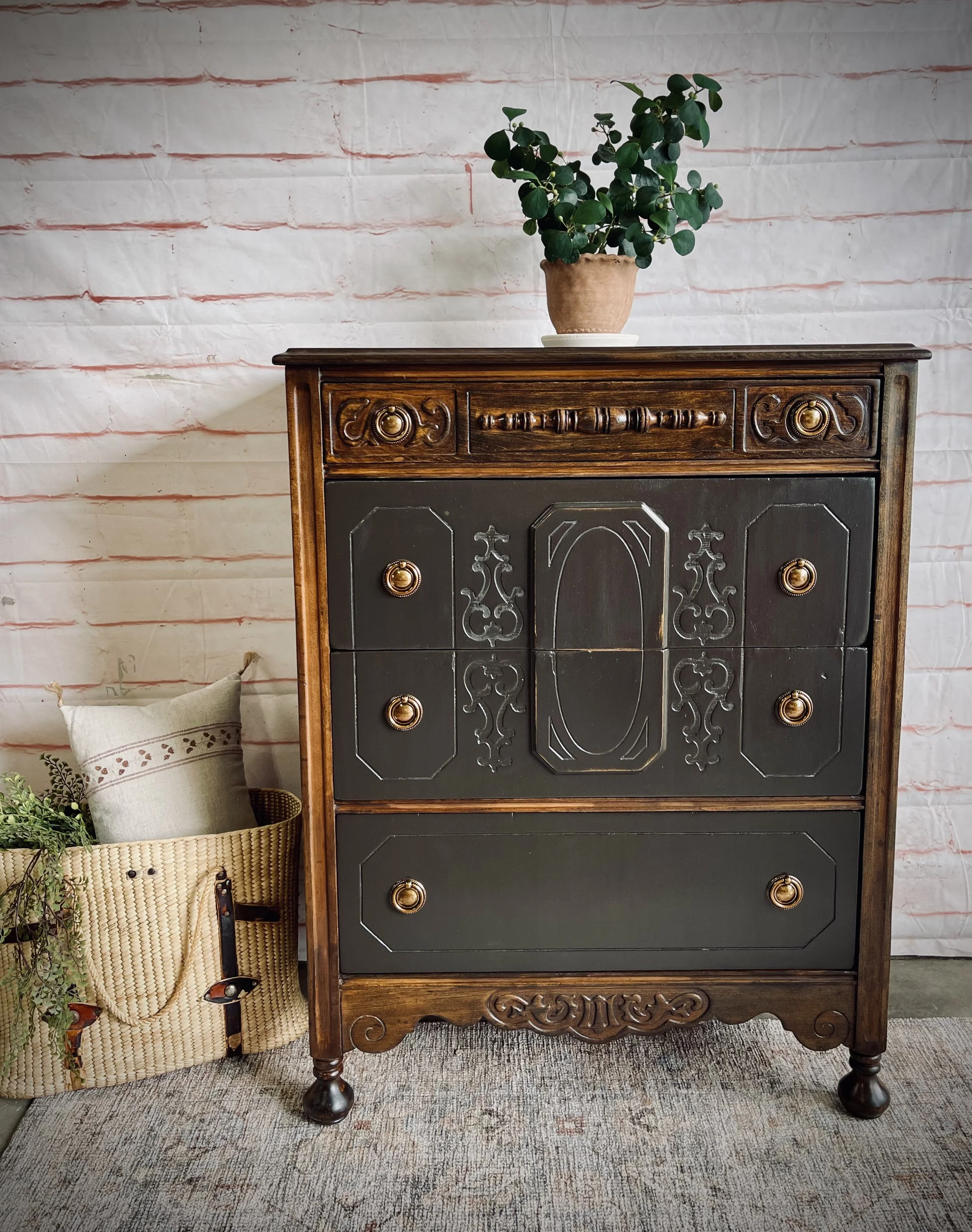 A vintage wooden dresser with black painted drawers and gold knobs, topped with a potted green plant, against a white brick wall with red accents. A basket with a blanket and plants is beside it.