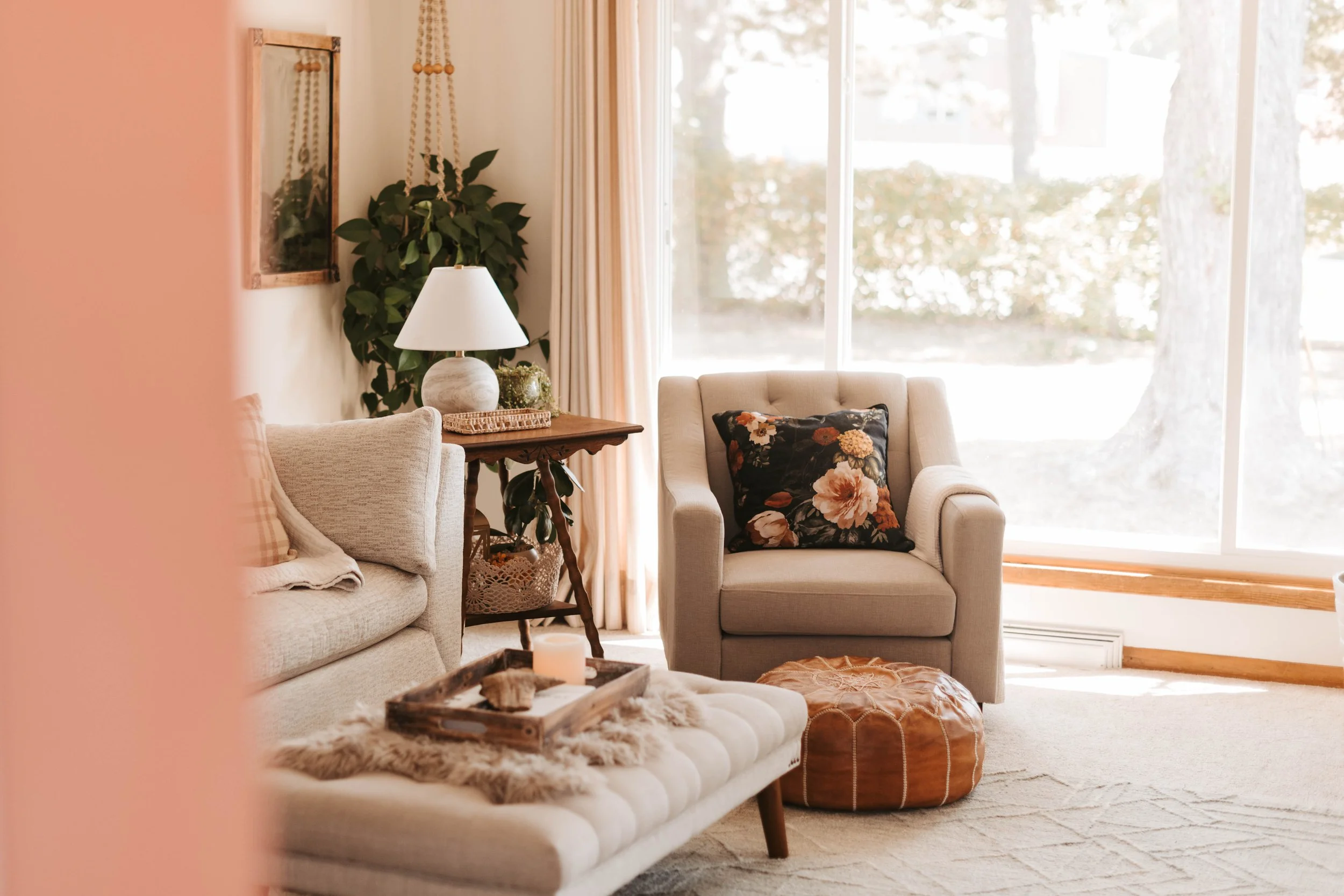 Cozy living room with beige armchair featuring a floral pillow, a padded bench with a tray and candle, a wooden side table with a lamp, a large window with curtains, and a leafy indoor plant.