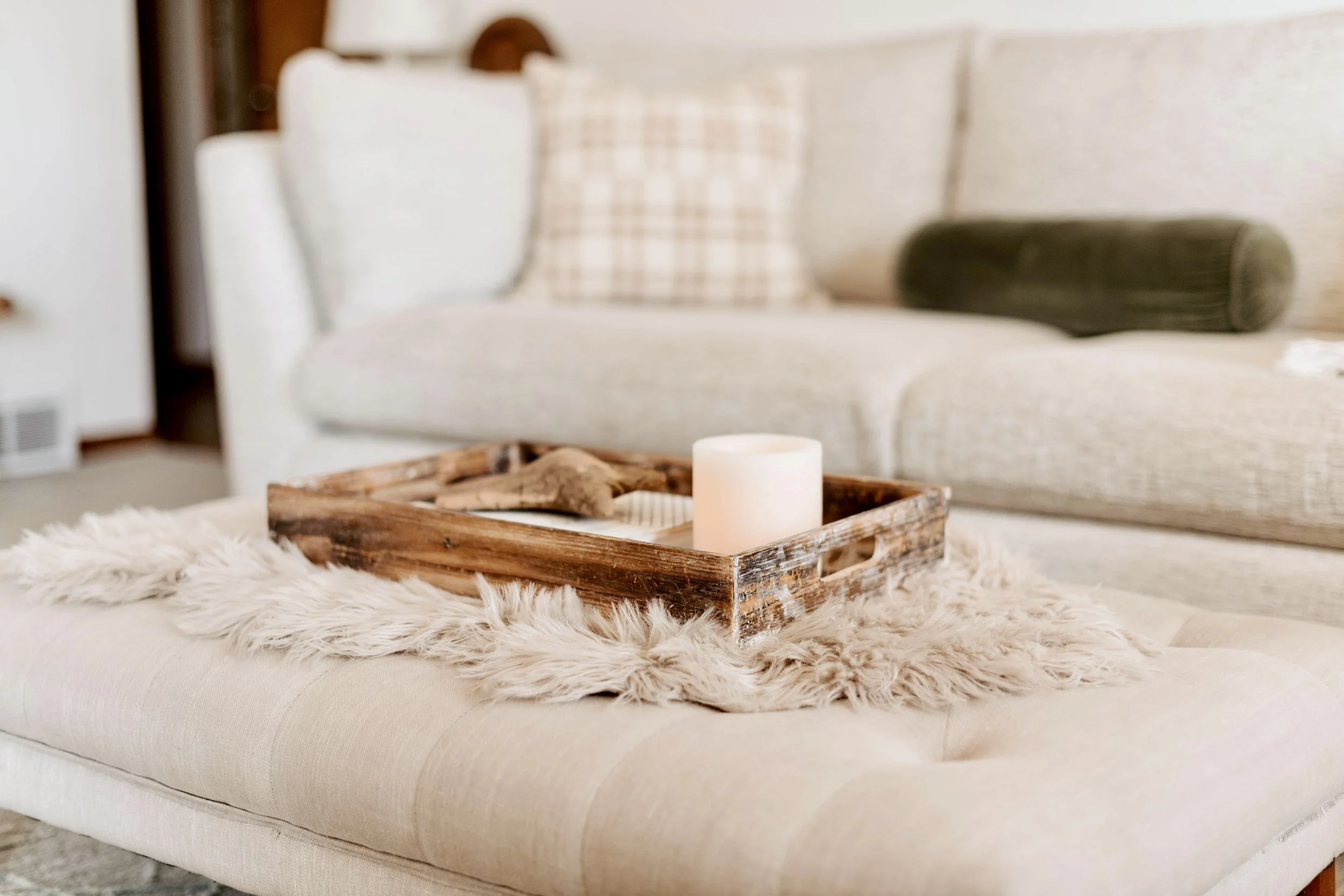Living room with a beige ottoman, a fluffy rug, a wooden tray, a candle, and a beige sofa with pillows in the background.