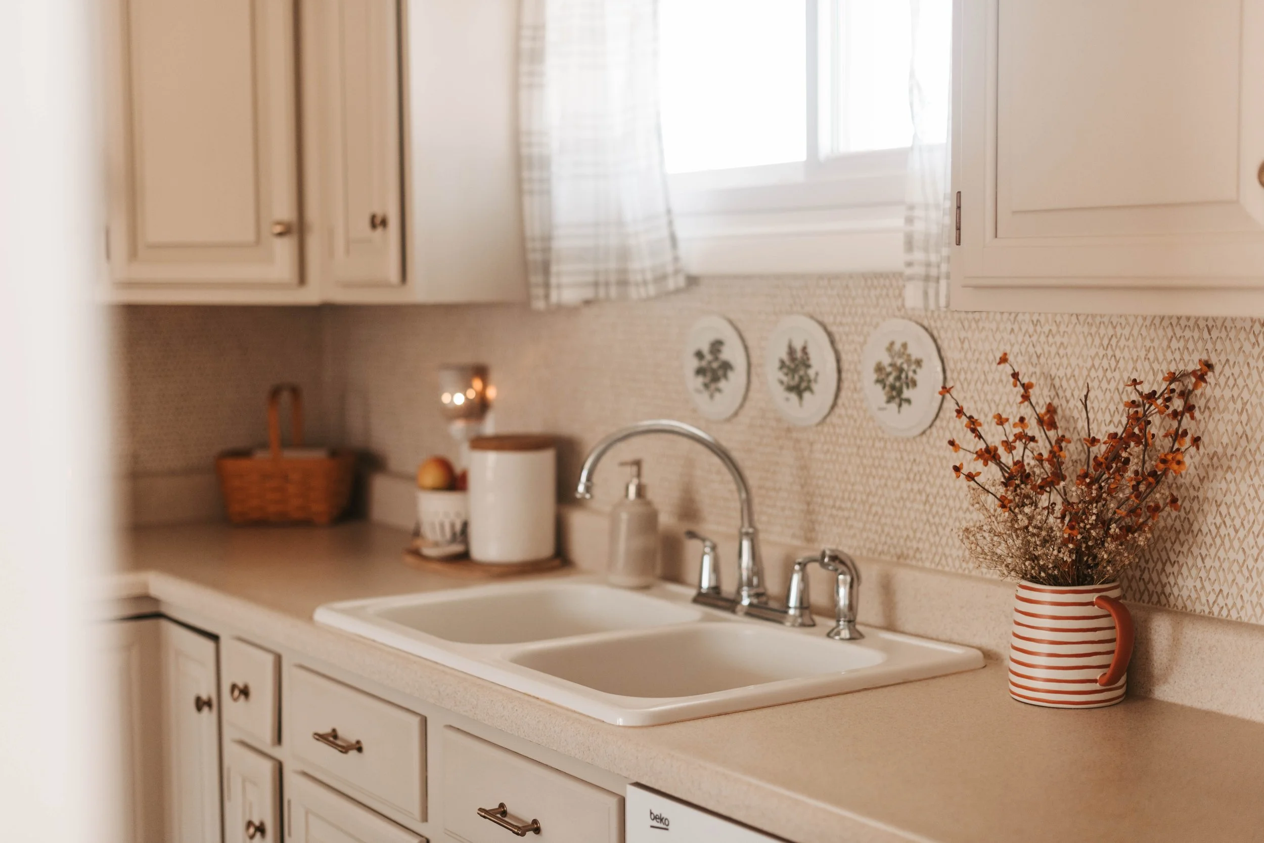 Kitchen countertop with a white farmhouse sink, a striped pitcher with dried flowers, soap dispenser, paper towel holder, and decorative plates with fruit and greenery on the wall