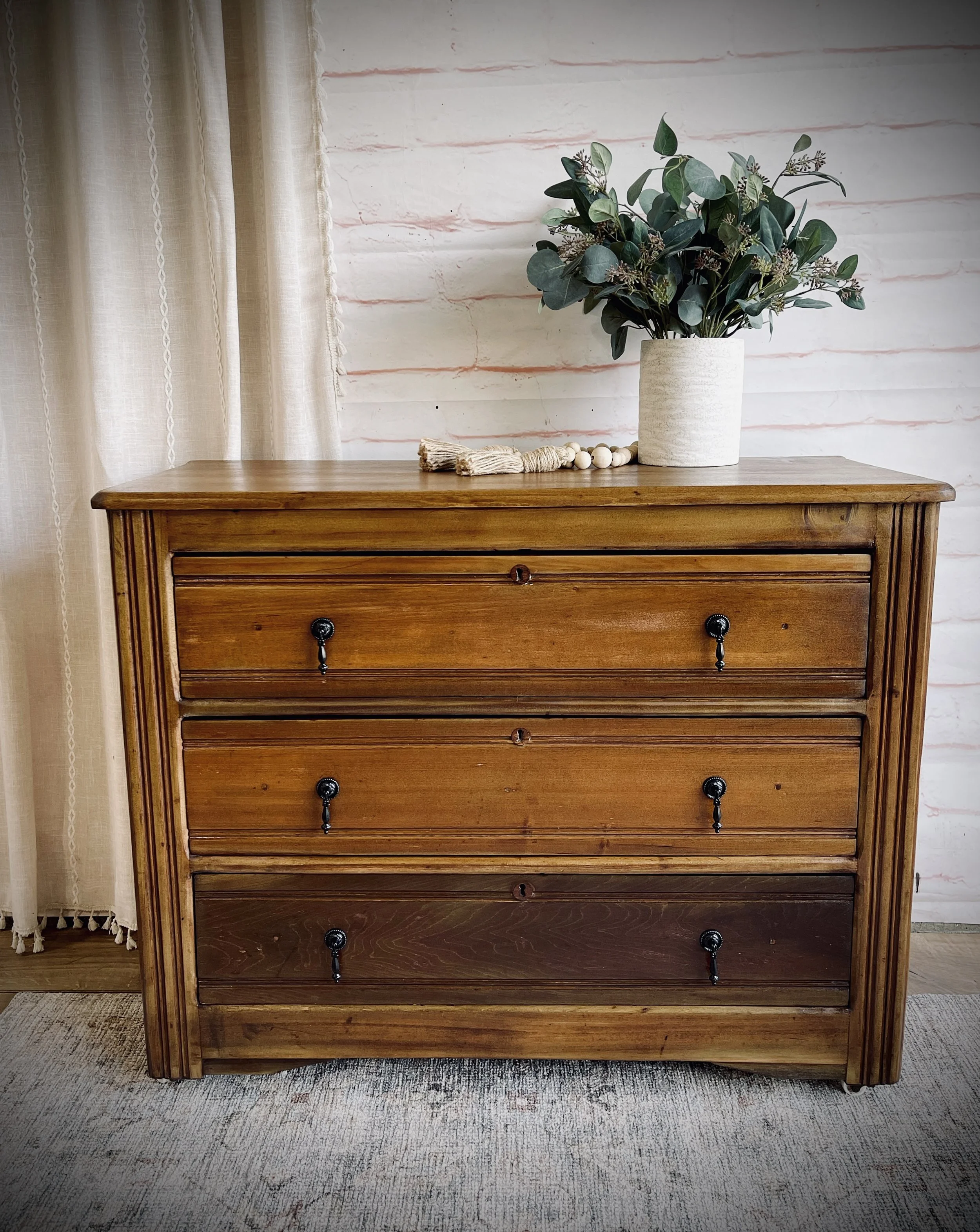 A wooden dresser with four drawers, decorated with a potted plant, a bead garland, and a cloth on top, set against a white brick wall with curtains to the left.