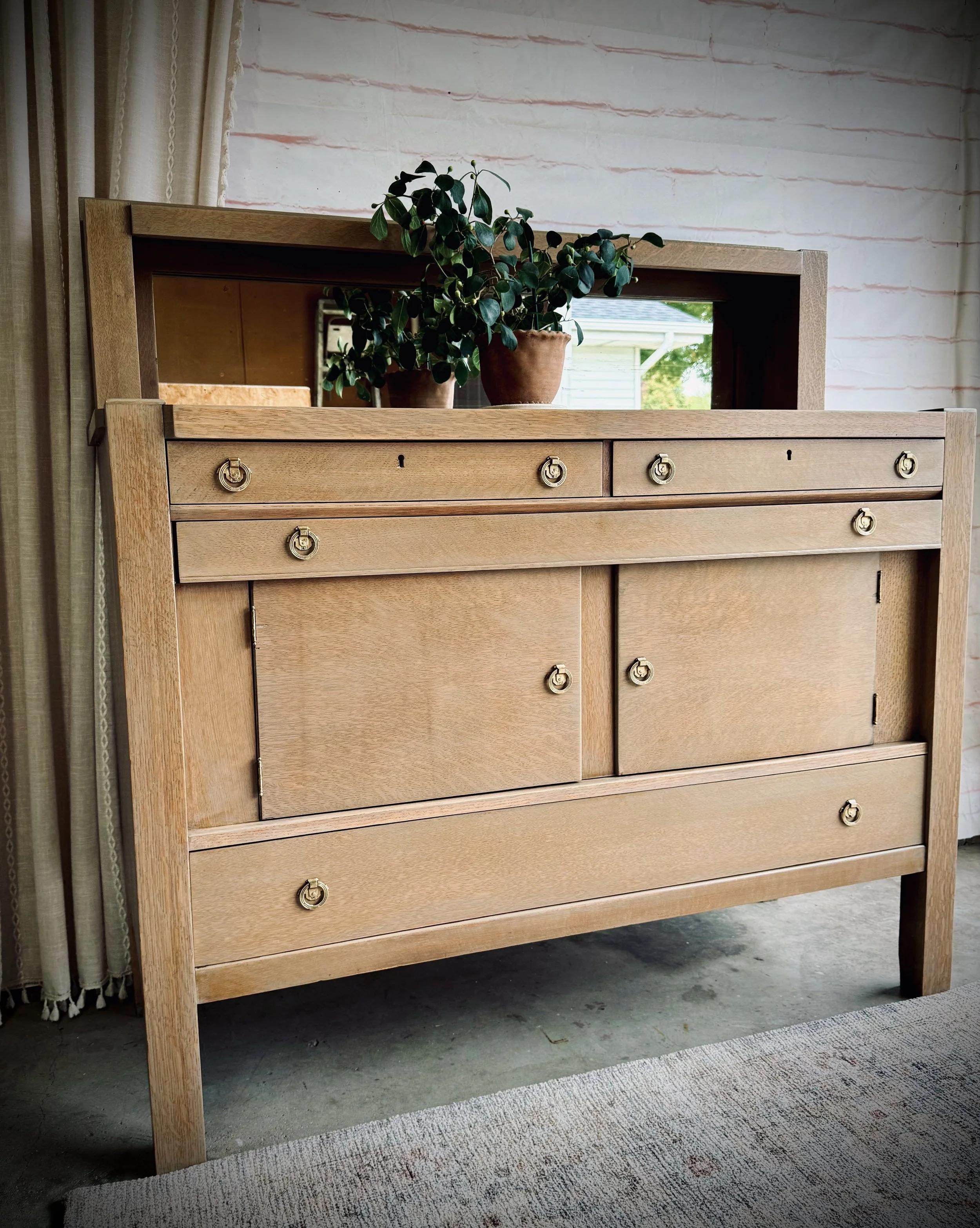 A wooden dresser with a mirror on top, decorated with two potted plants, standing on a concrete floor with a rug nearby, against a white brick wall.