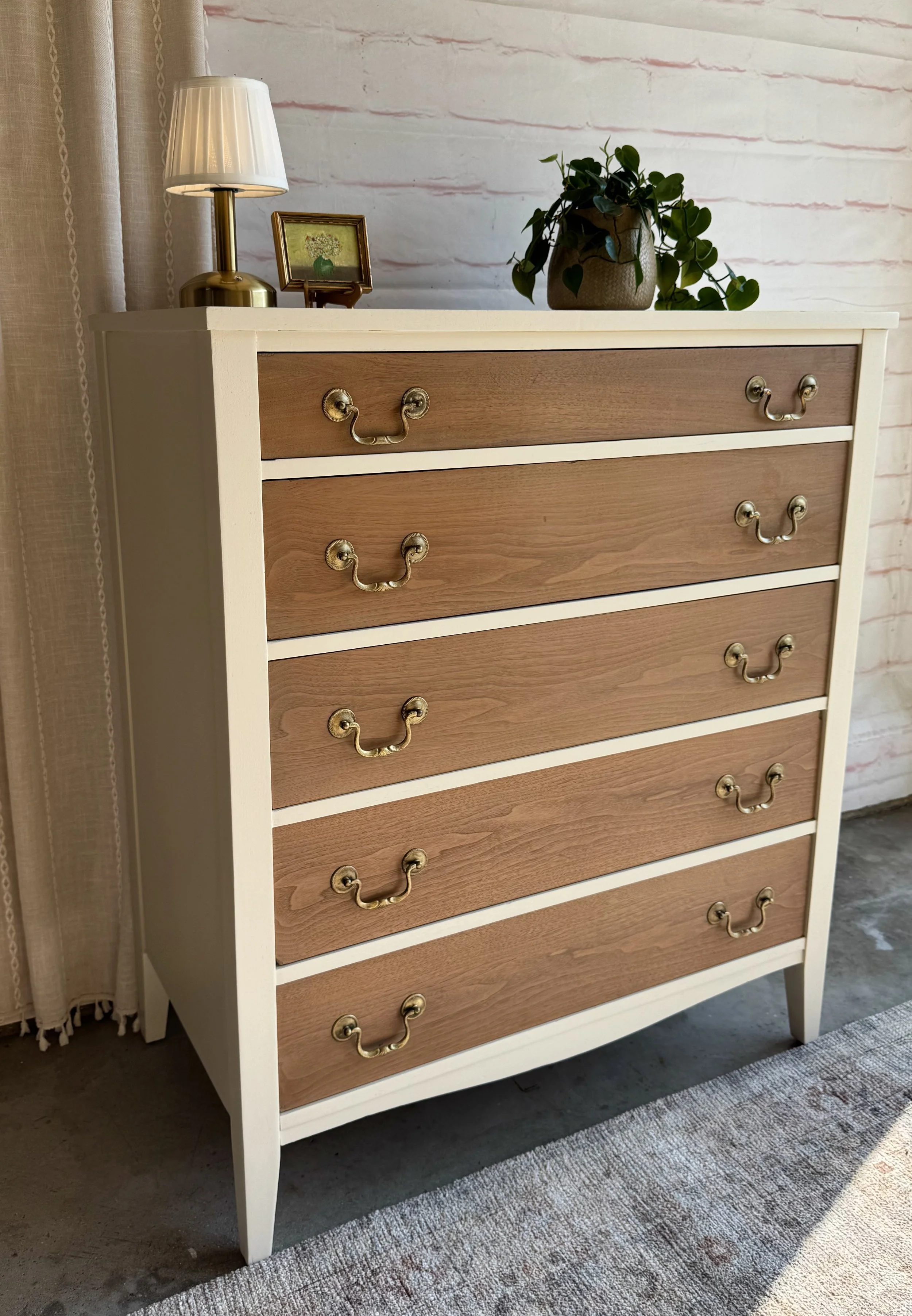 A dresser with six drawers, white frame, natural wood fronts, and brass handles. On top, there is a gold table lamp with a white shade, a small framed picture, and a potted plant with trailing green leaves.