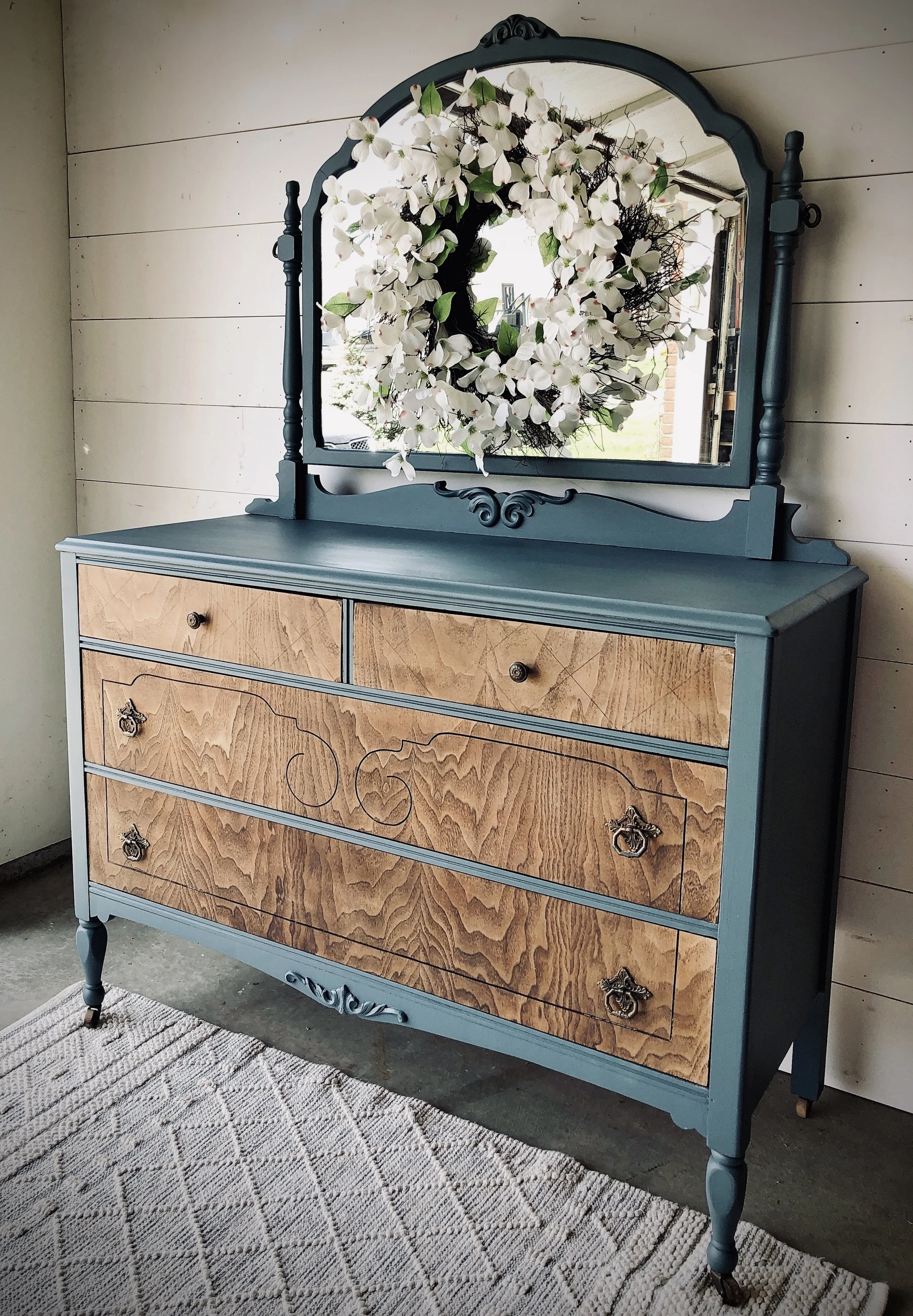 A blue vintage dresser with a mirror, decorated with a floral wreath, is placed against a white wooden wall and on a patterned rug.