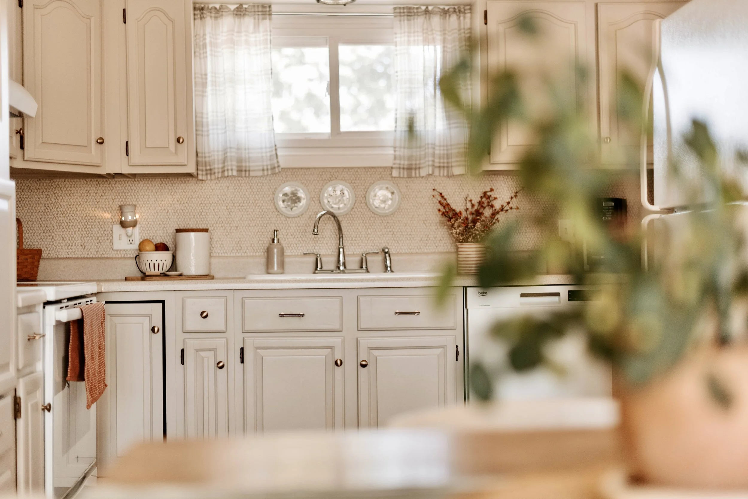 A cozy kitchen with beige cabinets, a window above the sink, and various kitchen items like a bowl of fruit, a paper towel, and a vase with flowers on the countertop. The kitchen has a warm, inviting atmosphere with natural light.