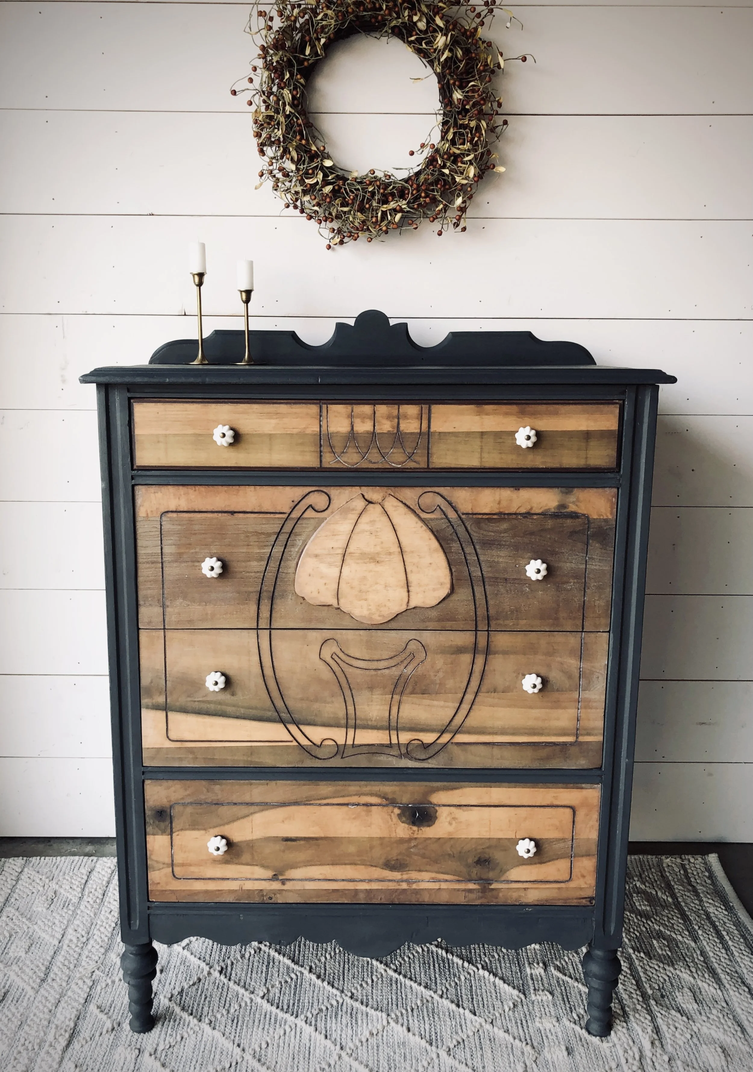 A wooden dresser with black trim and a floral design on the front, topped with two white candles on gold holders. Above it hangs a wreath made of twigs and small red berries. The background features a white paneled wall and a gray rug on the floor.