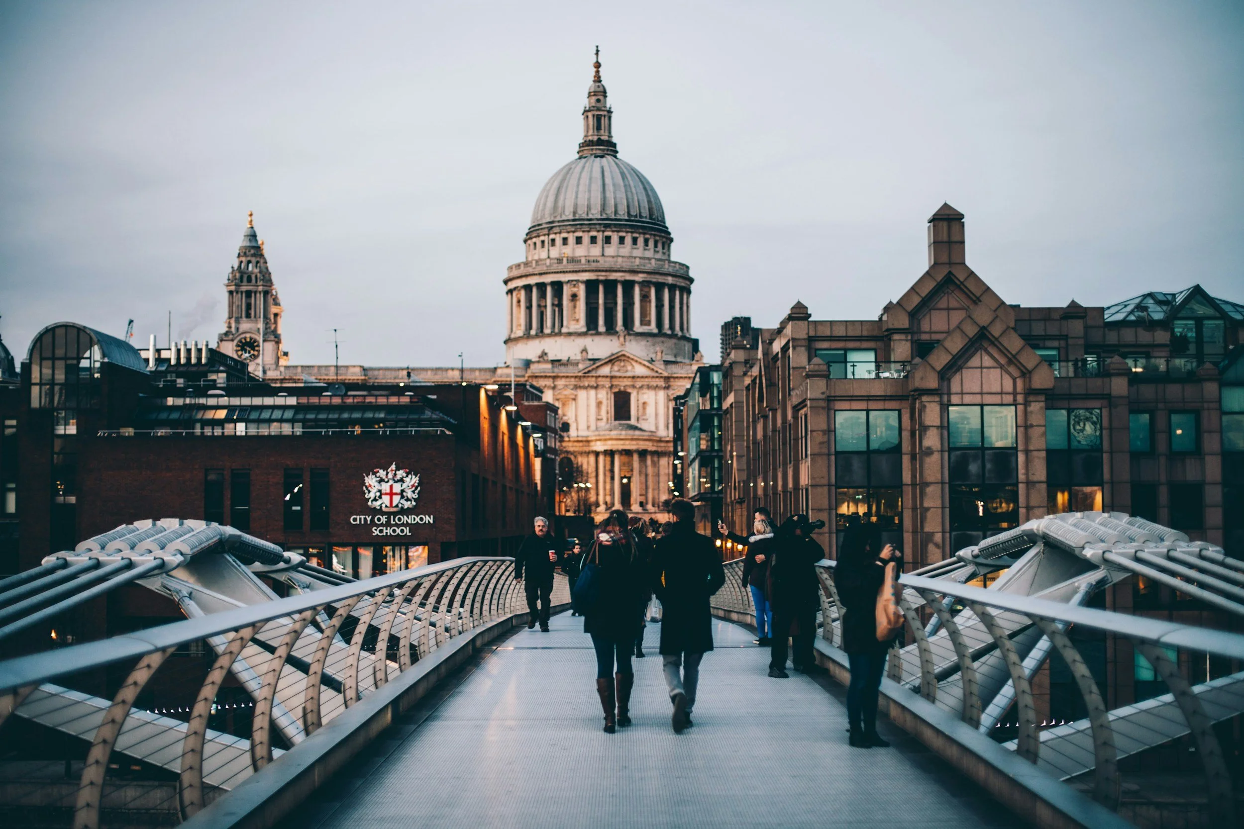 People walking on Millennium Bridge in London with St. Paul's Cathedral in the background, during dusk.