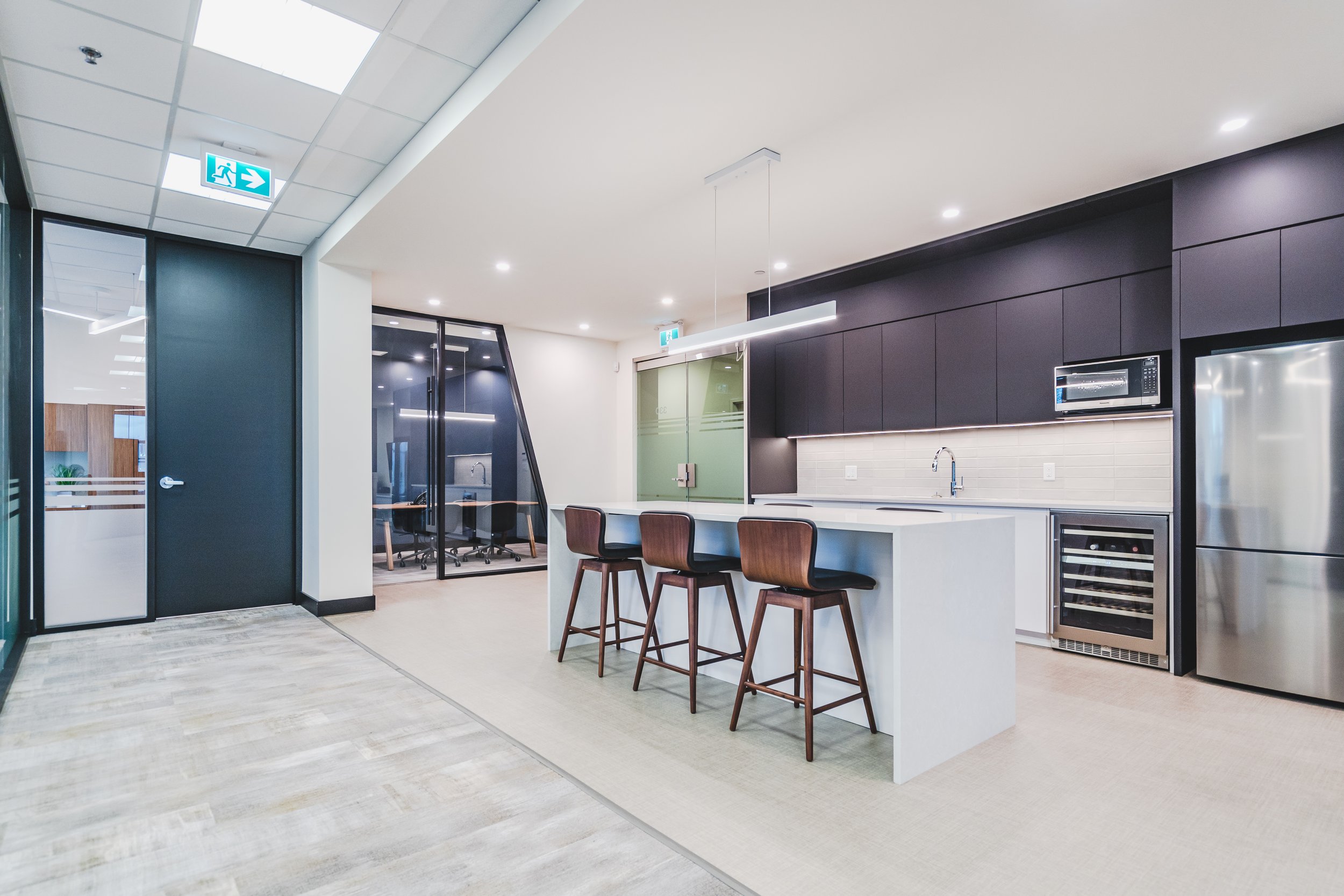 Modern office kitchen with a white island counter, three wooden bar stools, black cabinetry, stainless steel refrigerator, wine cooler, and microwave, with adjacent conference room with glass walls.