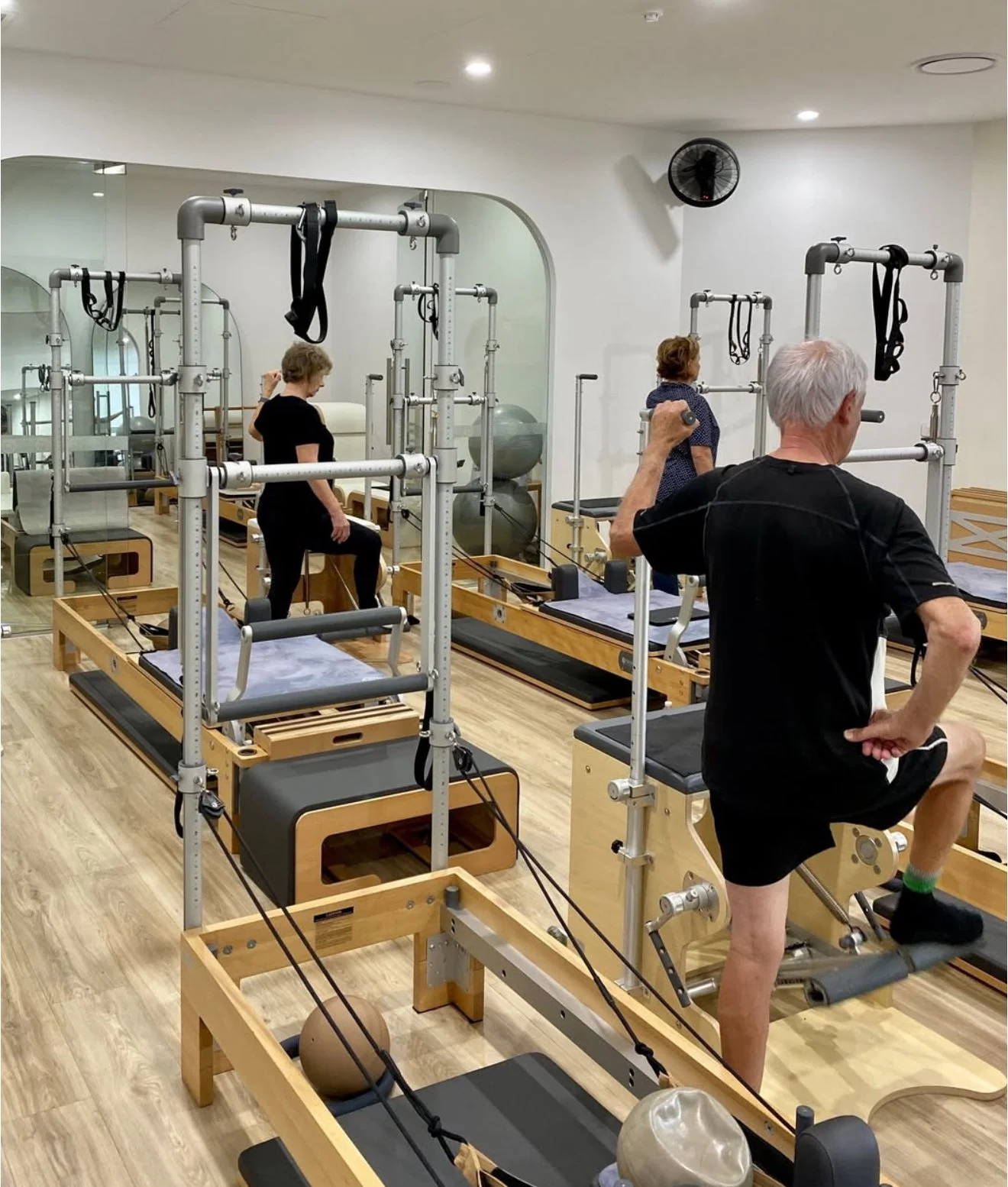 Senior adults and a middle-aged woman using Pilates reformer machines in a fitness studio with mirrors and wooden flooring.