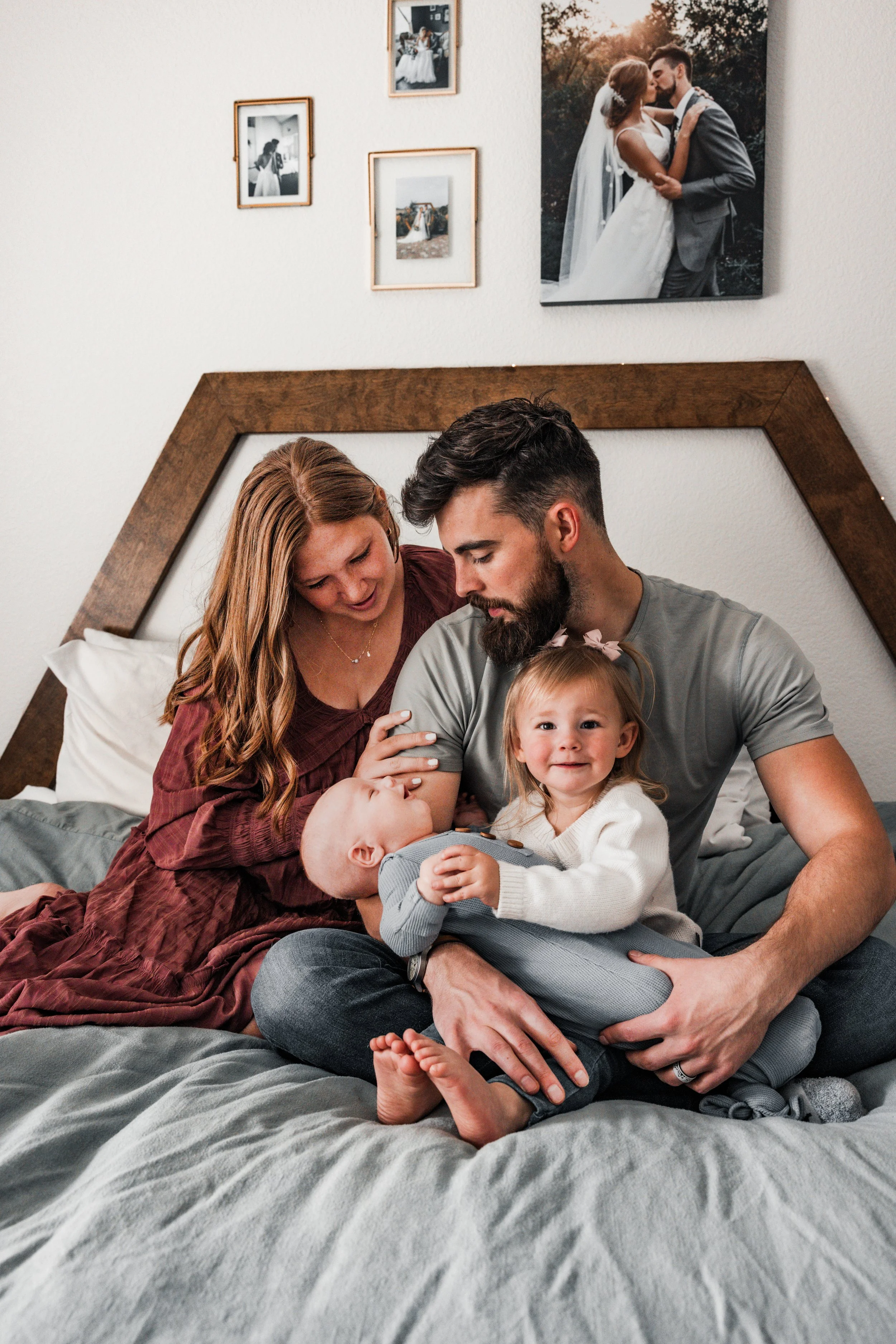 A family of four, including a woman with long red hair, a man with dark hair and a beard, a young girl, and a baby, sitting together on a bed in a bedroom, with framed photographs and a wedding picture of a couple on the wall behind them.