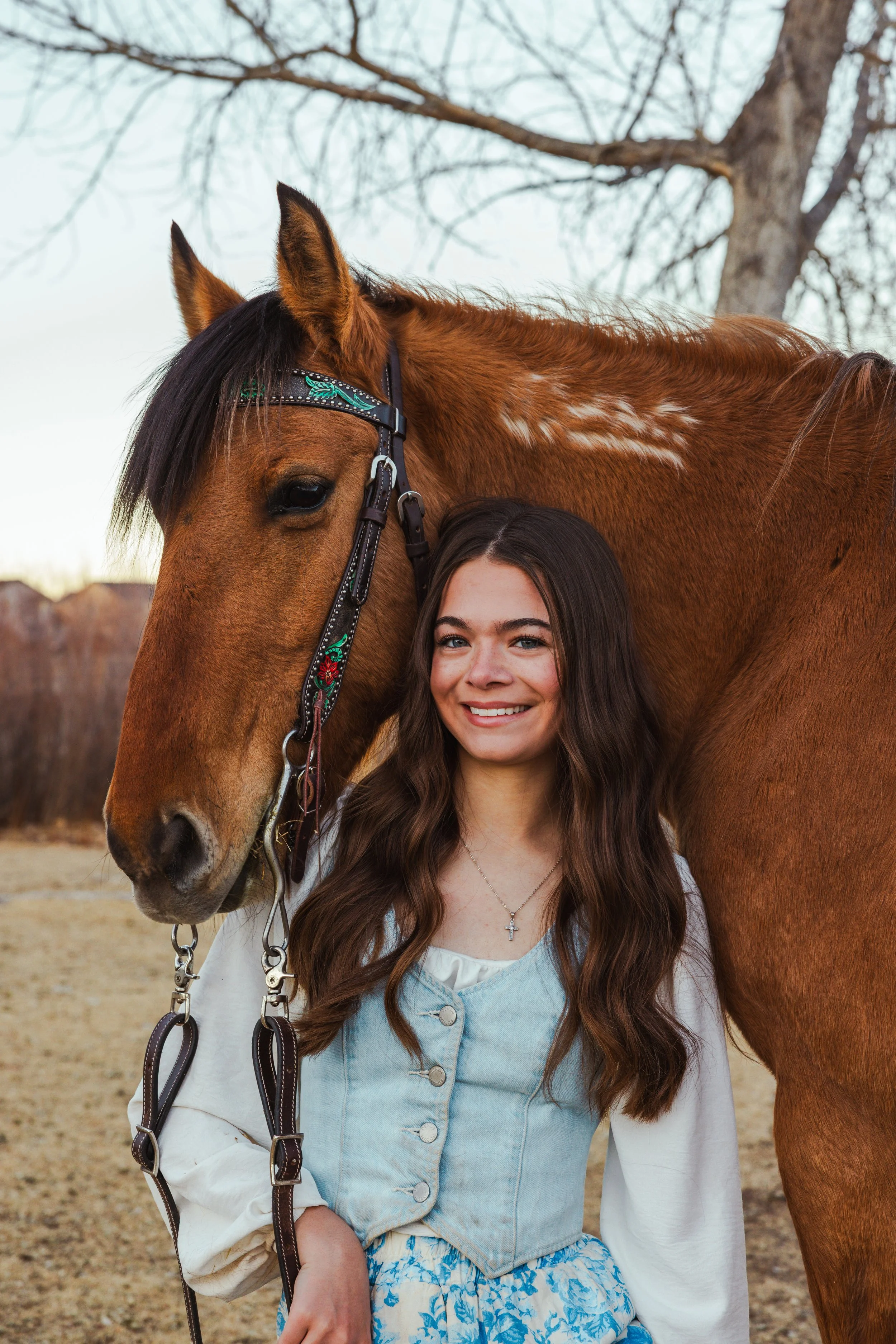 A young woman with long brown hair smiling, standing next to a brown horse with a patterned bridle in an outdoor setting with a leafless tree in the background.