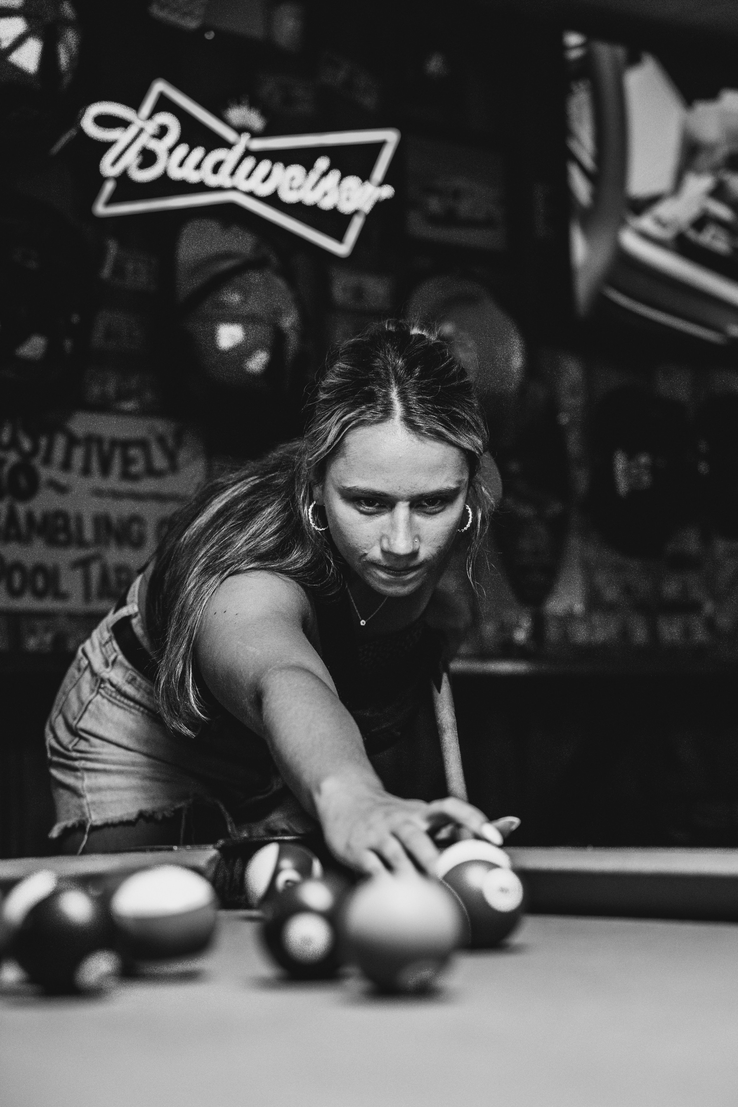 A woman playing pool at a bar or pool hall, focused as she lines up her shot, with billiard balls on the table, neon signs and posters on the wall in the background.