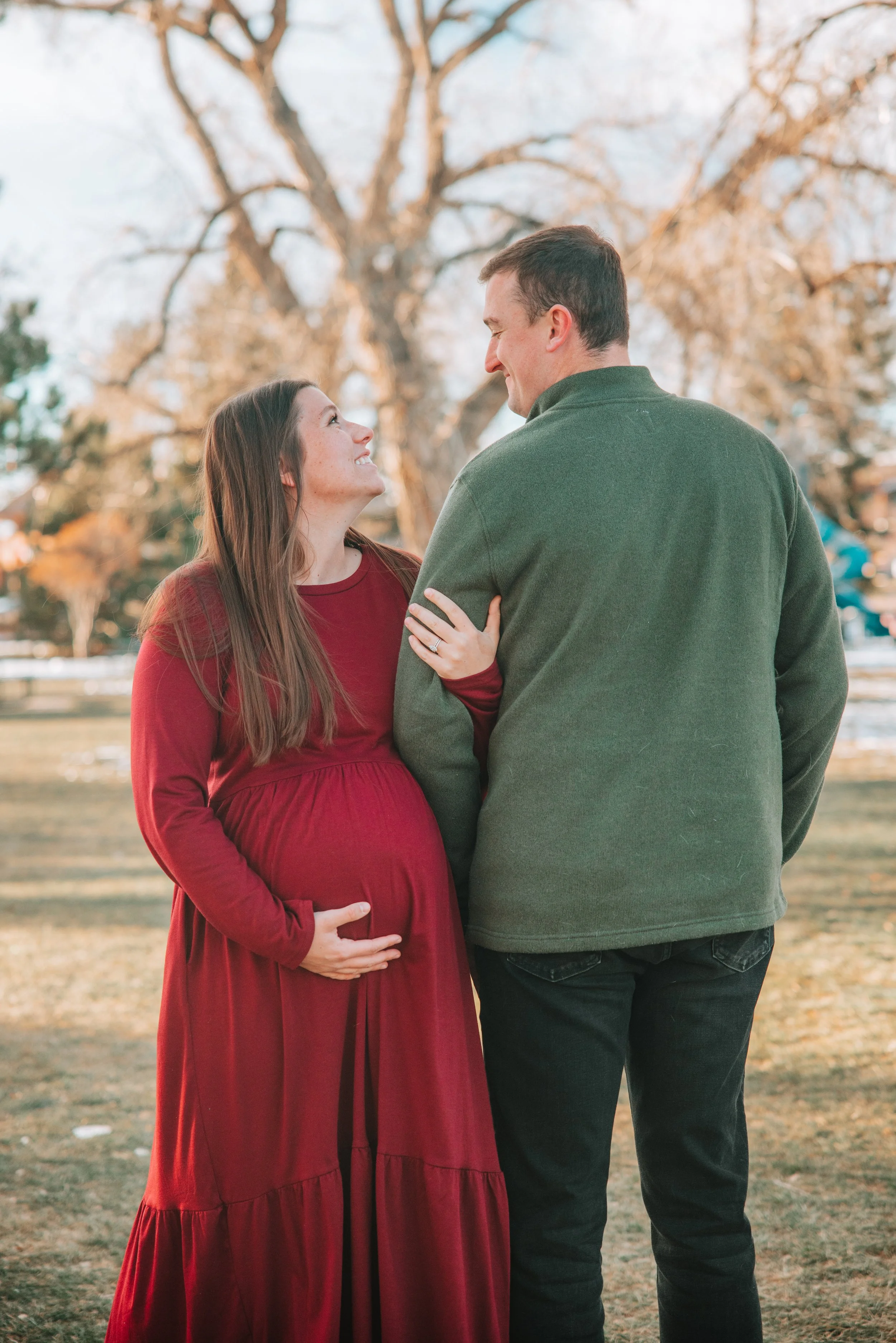 A pregnant woman with long brown hair wearing a red dress smiling at a man and holding her belly, standing outdoors in a park with trees in autumn foliage. The man, with short dark hair and wearing a green jacket, is smiling back at her.