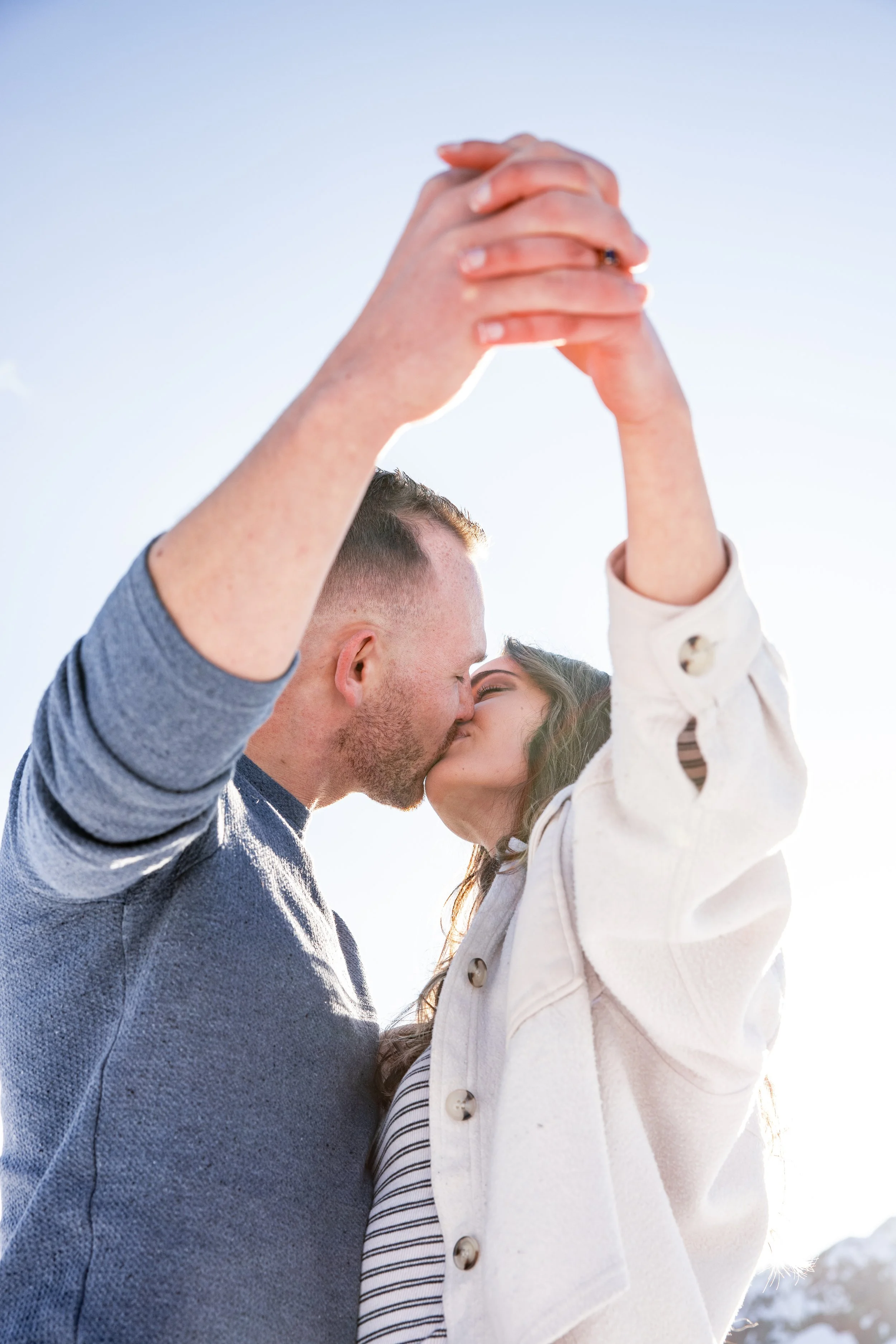 A couple is holding hands above their heads and kissing outdoors with blue sky and snow in background.