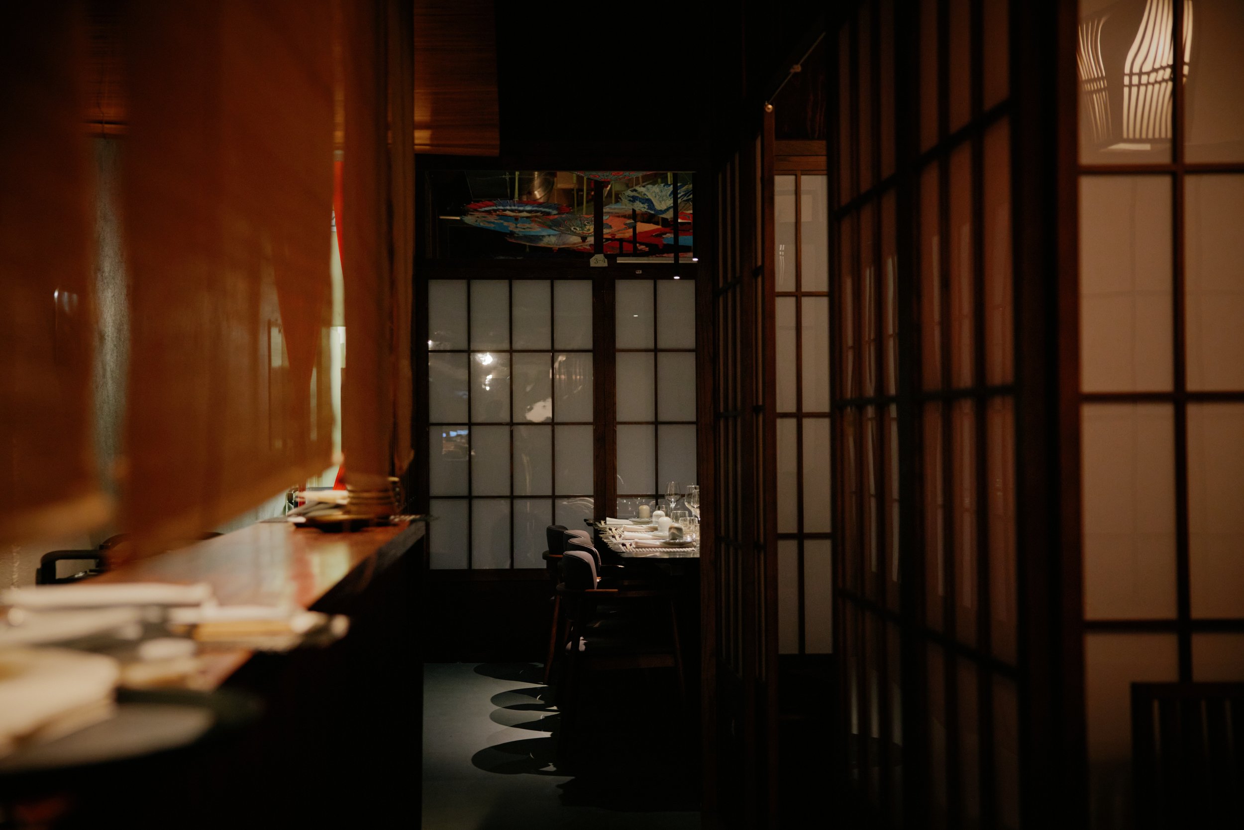 Interior of a dimly lit restaurant with wooden partition panels and a long table set with plates, glasses, and silverware.