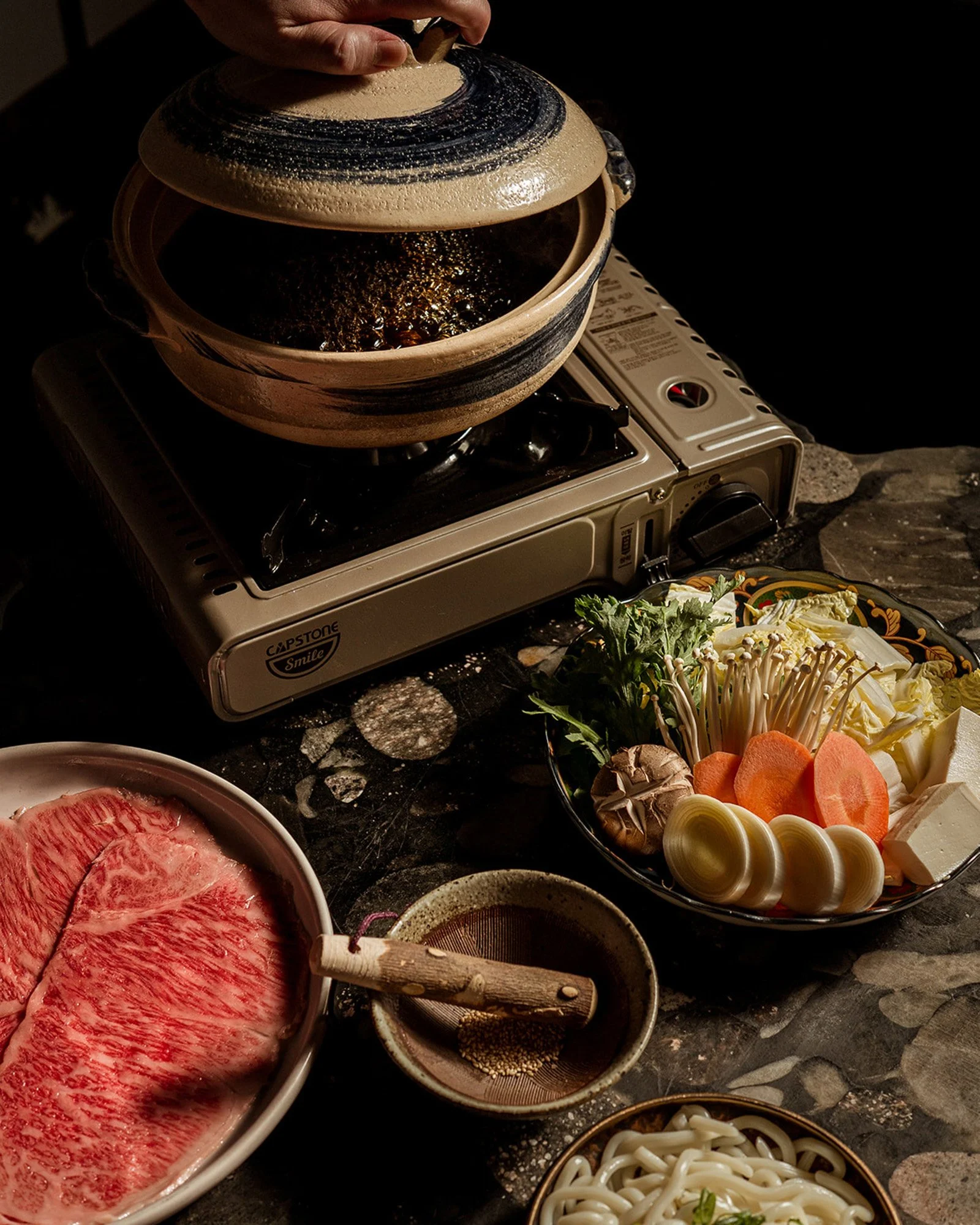 A hot pot being cooked on a portable stove, surrounded by fresh ingredients including sliced beef, vegetables, and noodles.