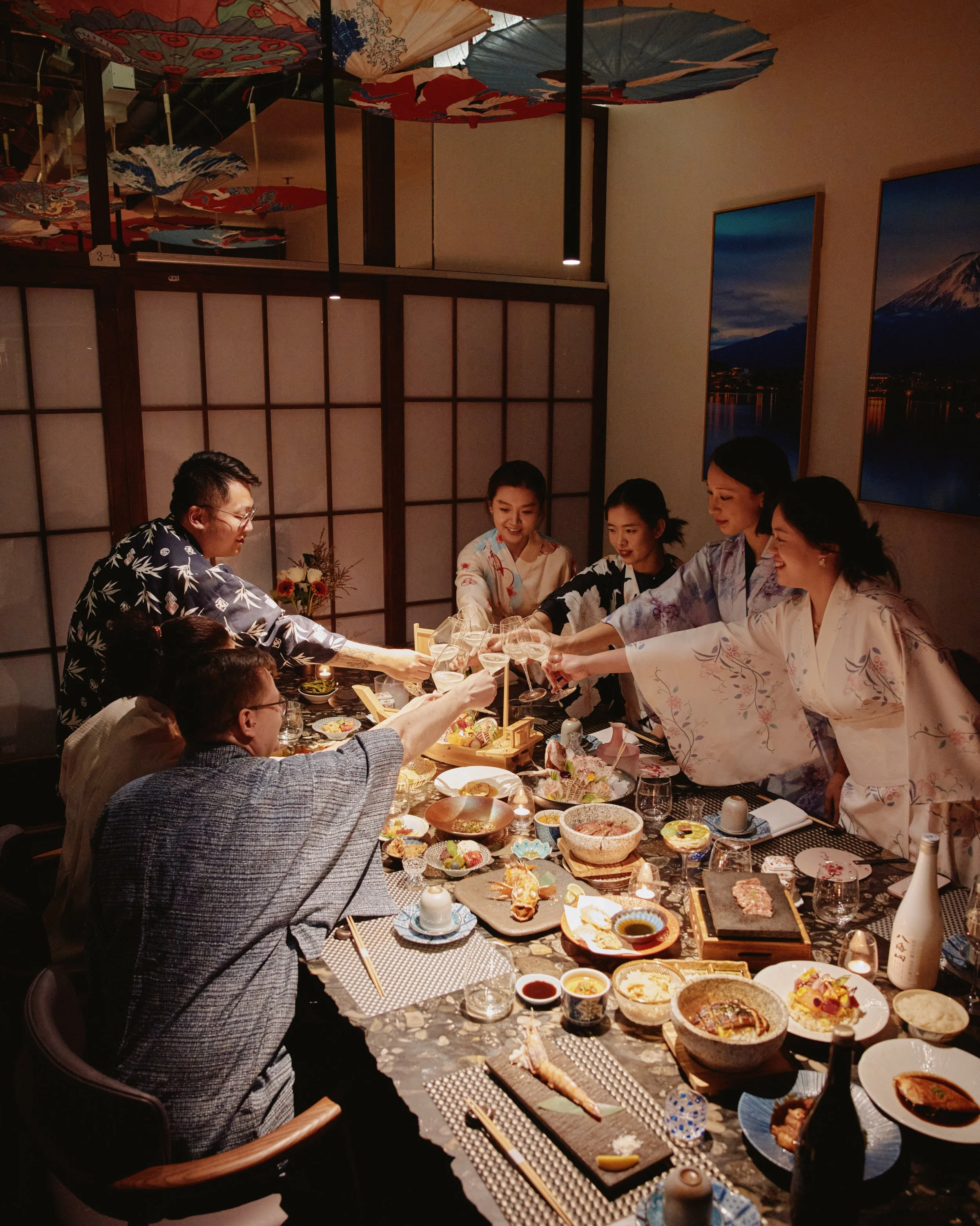 People in traditional Japanese kimonos enjoying a dinner gathering around a long table filled with various Japanese dishes, drinks, and tableware in a cozy decorated room with umbrellas hanging from the ceiling.