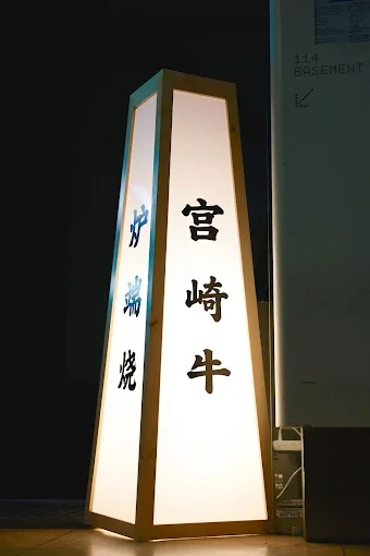 A tall, illuminated sign with Japanese characters in black on a white background, with a dark frame around the edges, signaling the restaurant entrance.