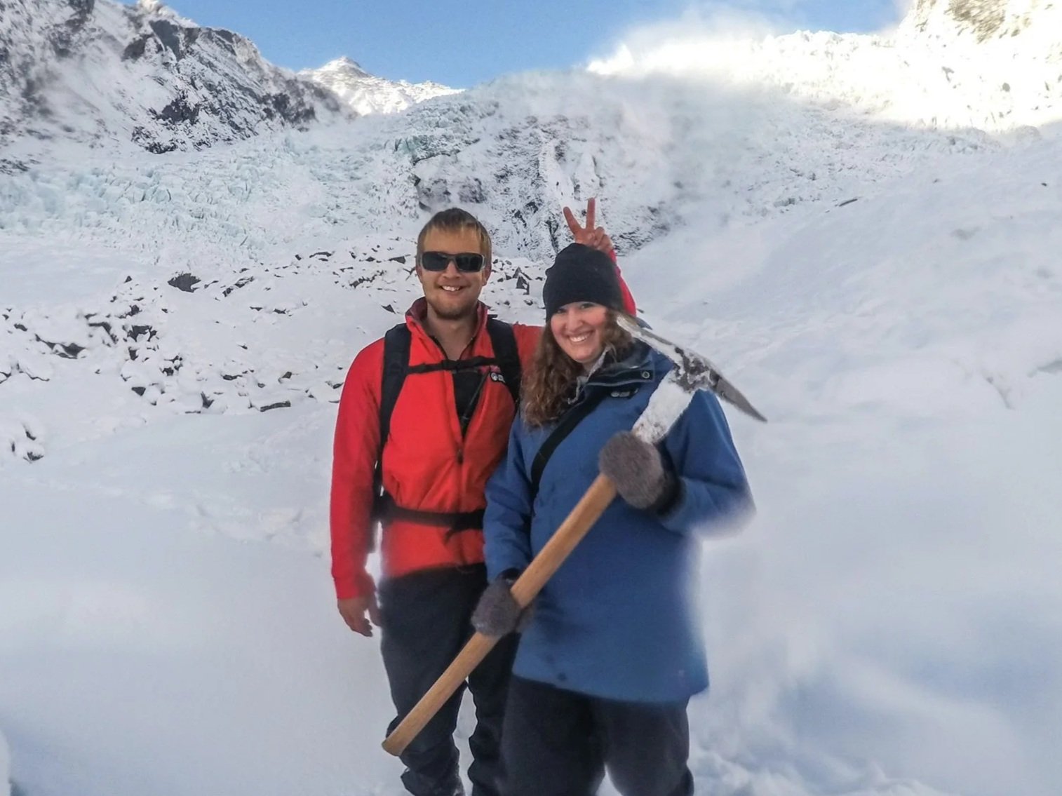 Two people in winter clothing standing in a snowy landscape, one woman holding an ice axe and the man making a peace sign behind her, with snow-covered mountains in the background.