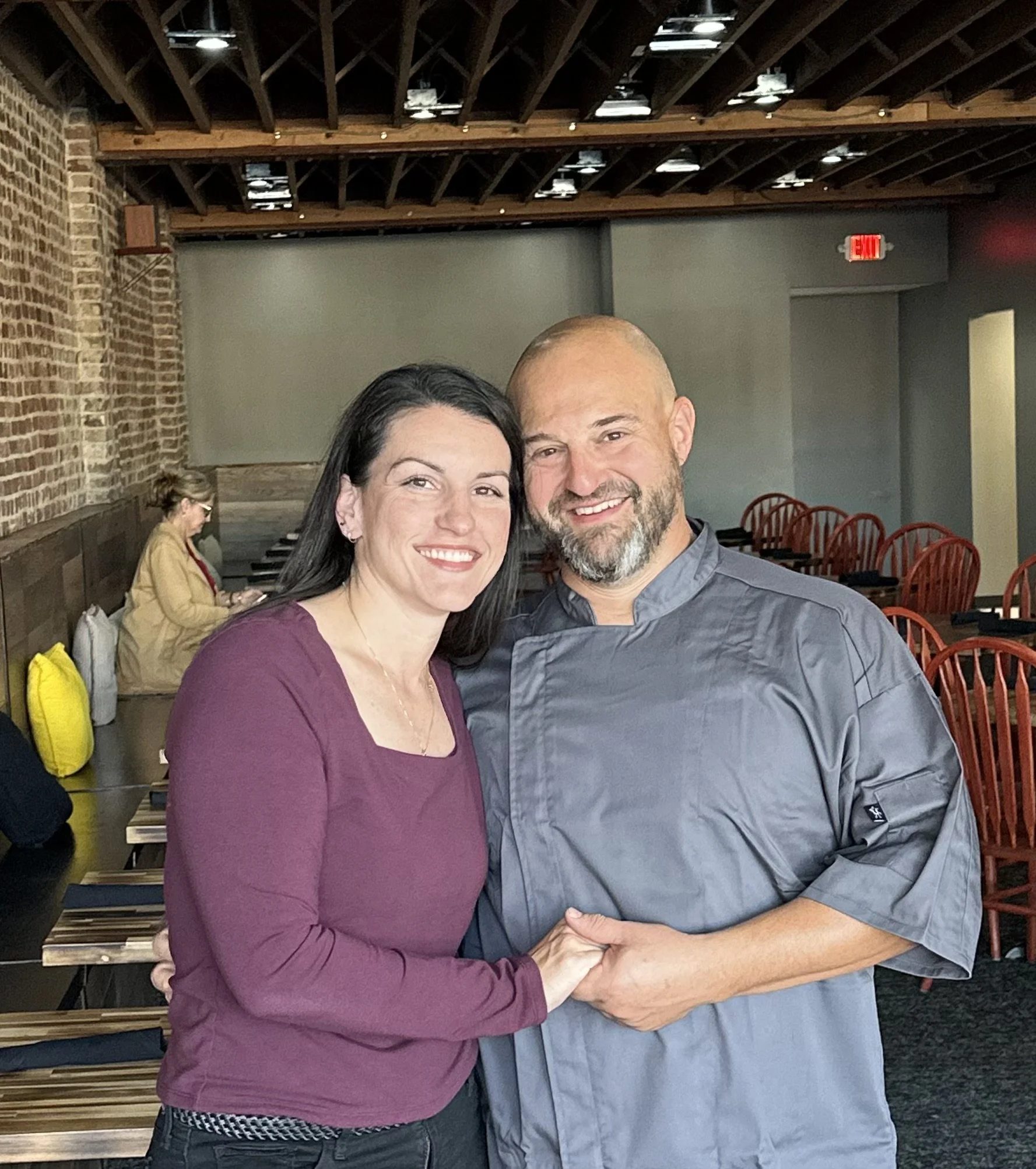 A woman and a man, one in a purple long-sleeve shirt and the other in a gray chef jacket, smiling and holding hands in a restaurant with a brick wall and red chairs in the background.