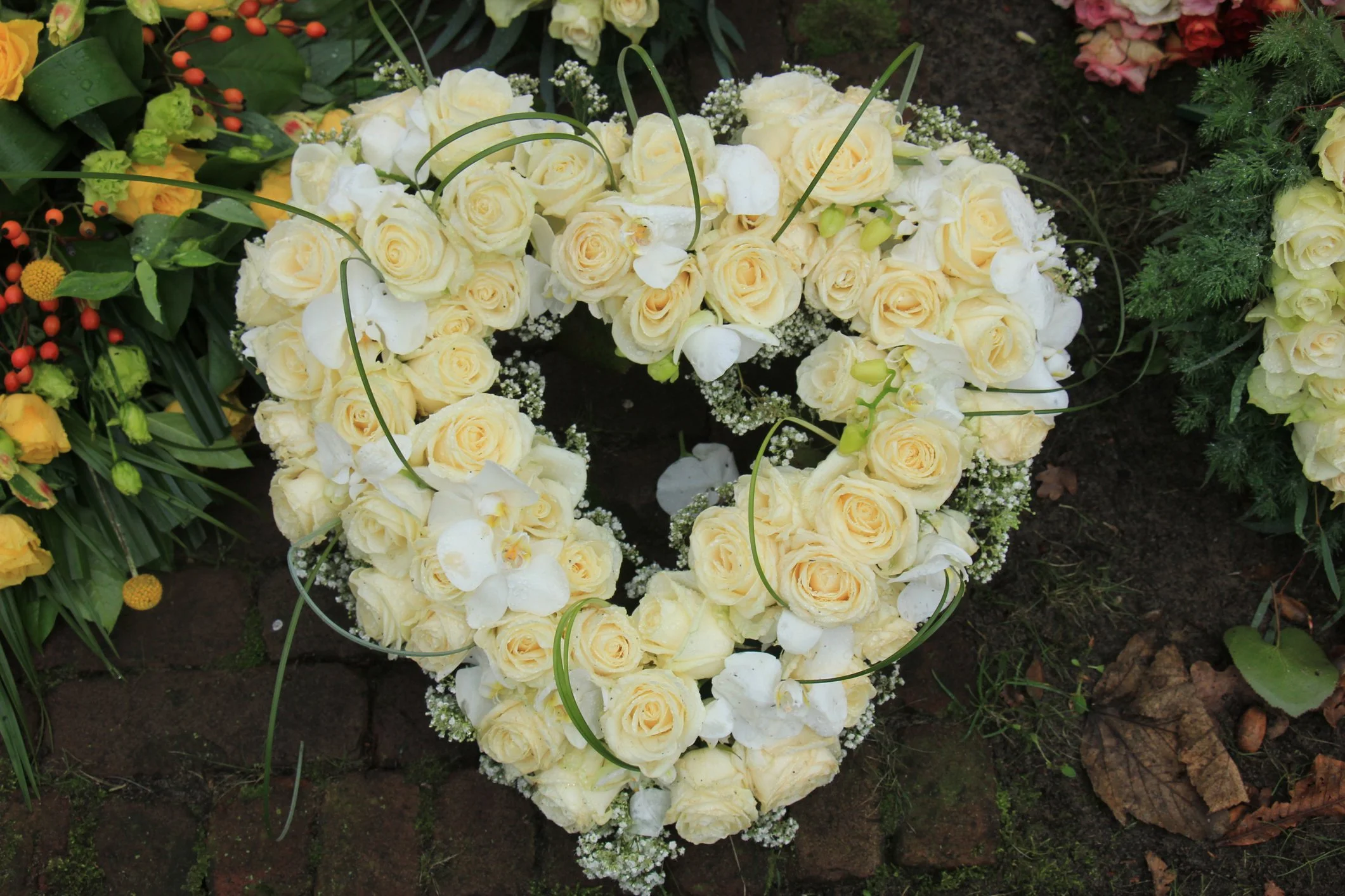 A funeral flower arrangement of pink, red, white, and cream roses, lilies, and greenery on top of a white casket, with a cloudy sky in the background.