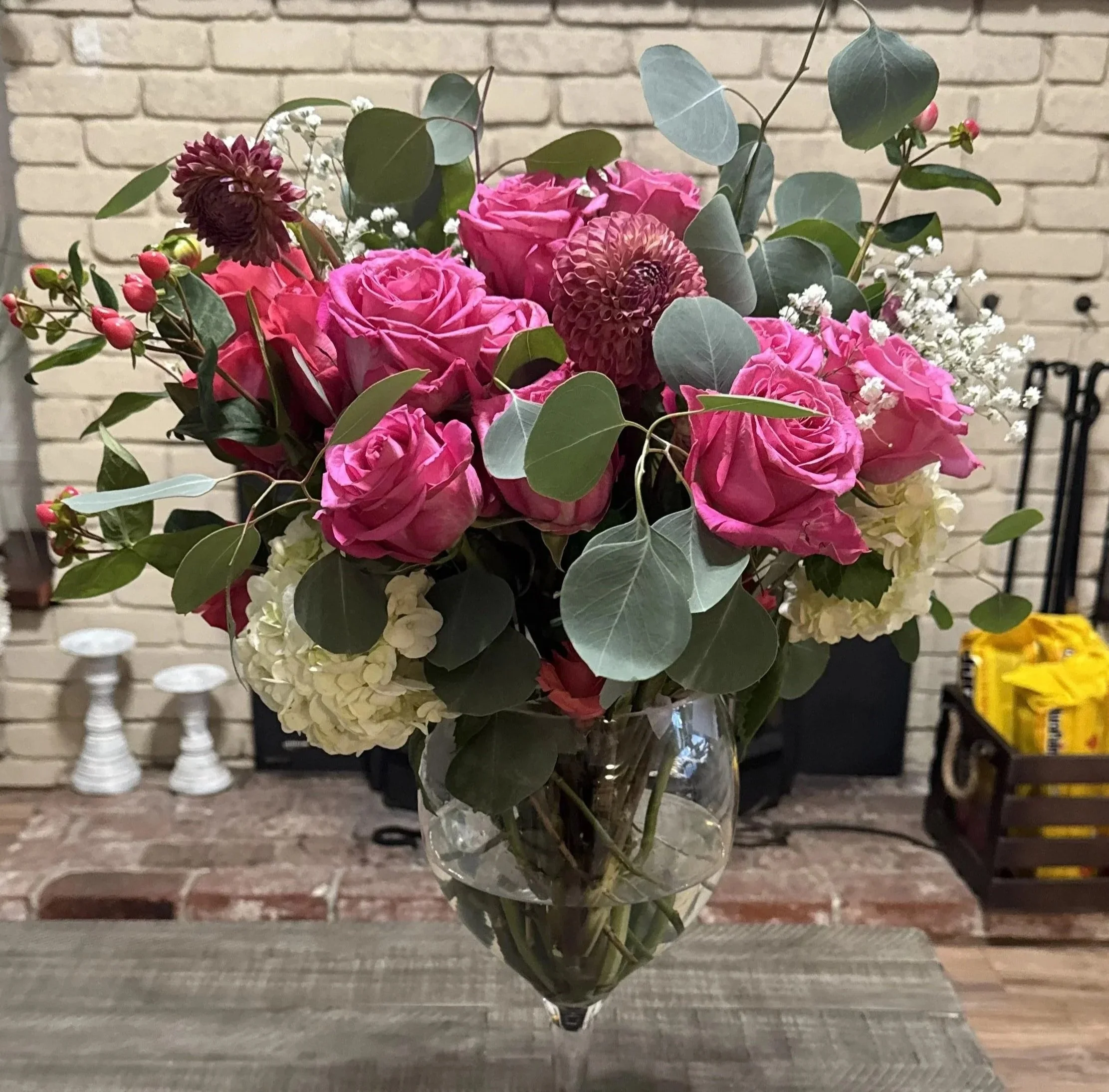 A bouquet of pink roses, white hydrangeas, and assorted greenery in a glass vase placed on a wooden table in front of a brick fireplace.