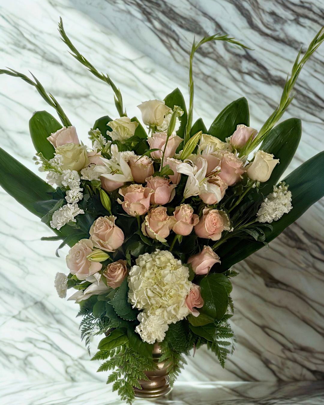 A white floral wreath made of cream-colored roses, white orchids, and baby's breath, resting on the ground with some surrounding greenery and other flowers.