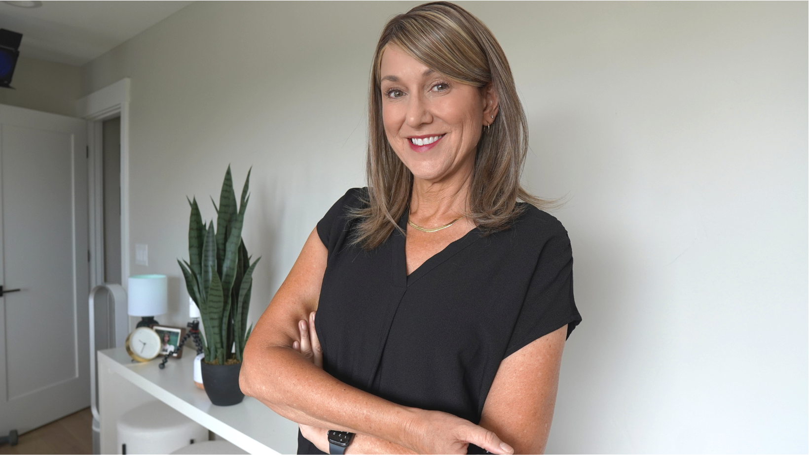 A smiling woman with shoulder-length brown hair, wearing a black top, standing with her arms crossed in a modern room with a white wall, a white console table, a tall green snake plant, a white lamp, a clock, and a framed photo.