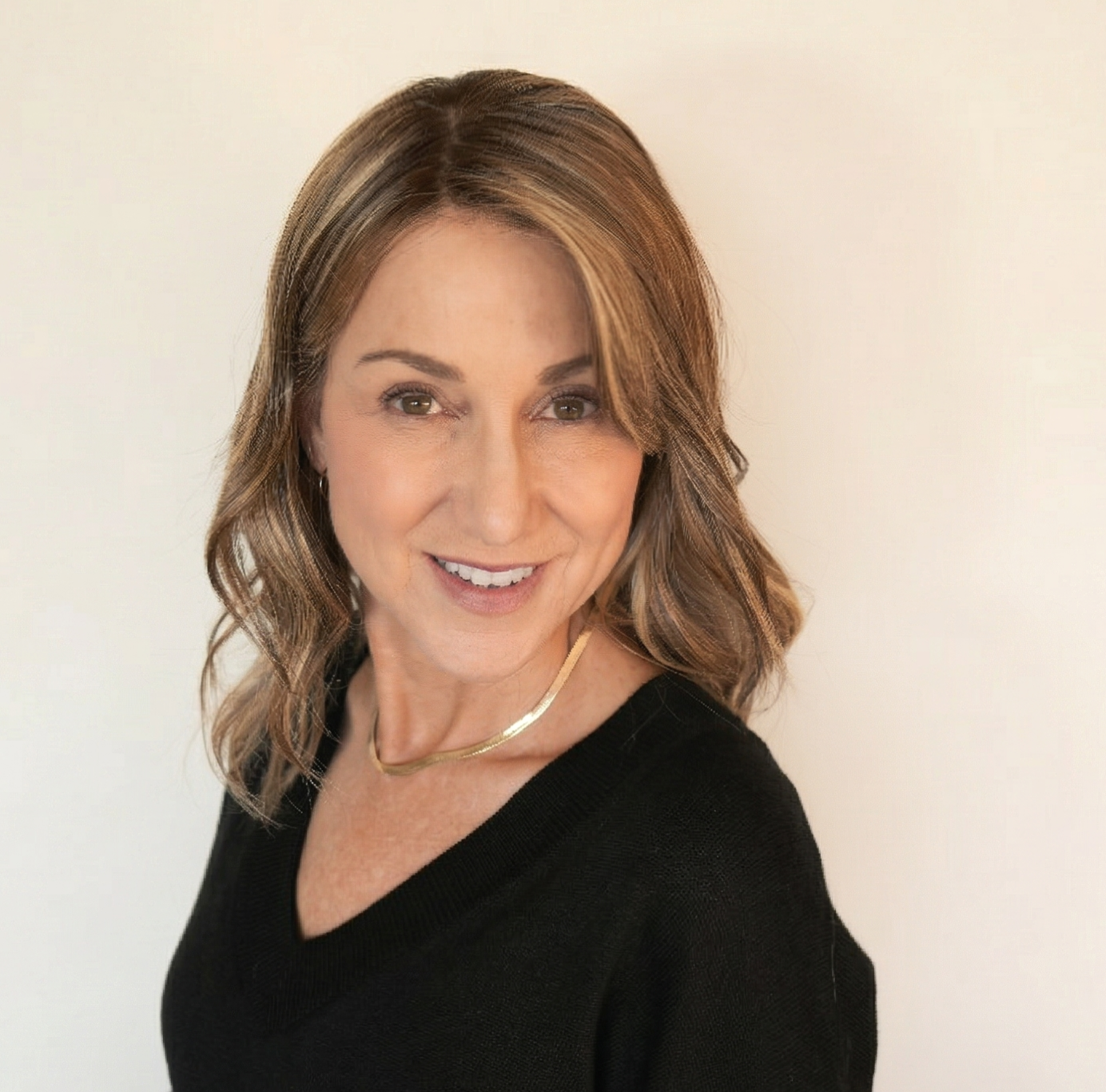 Portrait of a woman with shoulder-length brown hair with highlights, wearing a black top and a gold necklace, smiling at the camera against a plain off-white background.