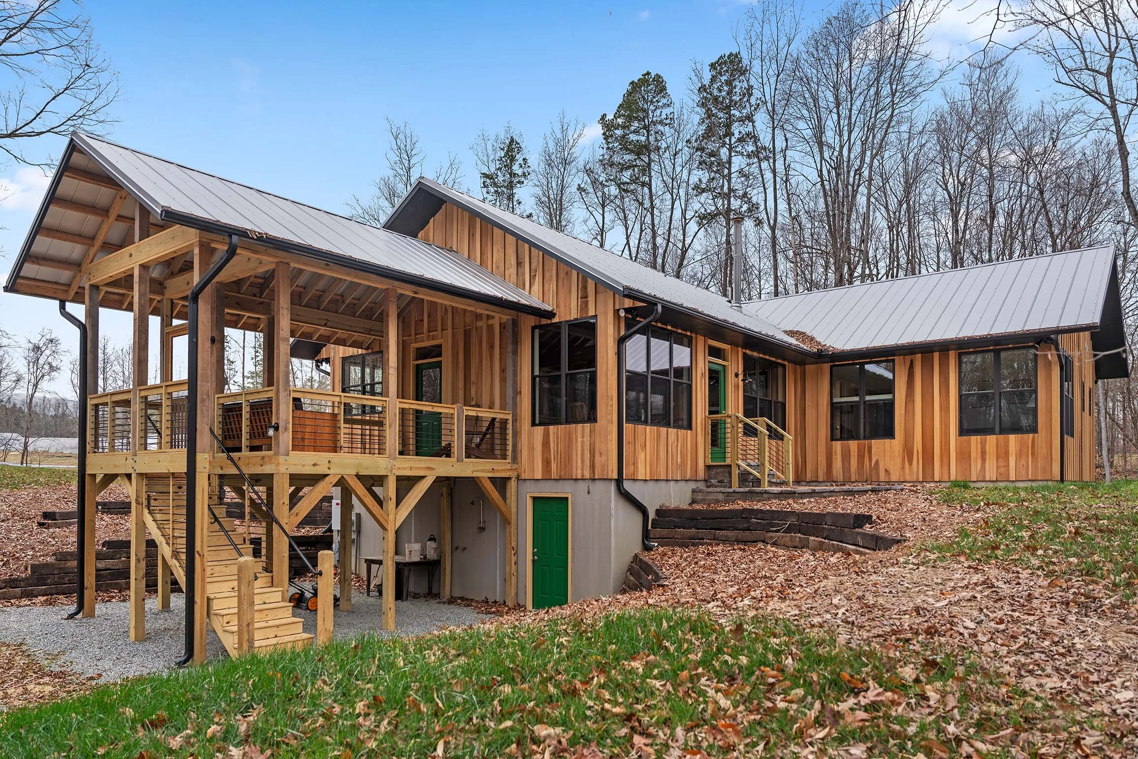 Newly built wooden house with a metal roof, multiple windows, and a balcony, surrounded by trees and a yard with fallen leaves.