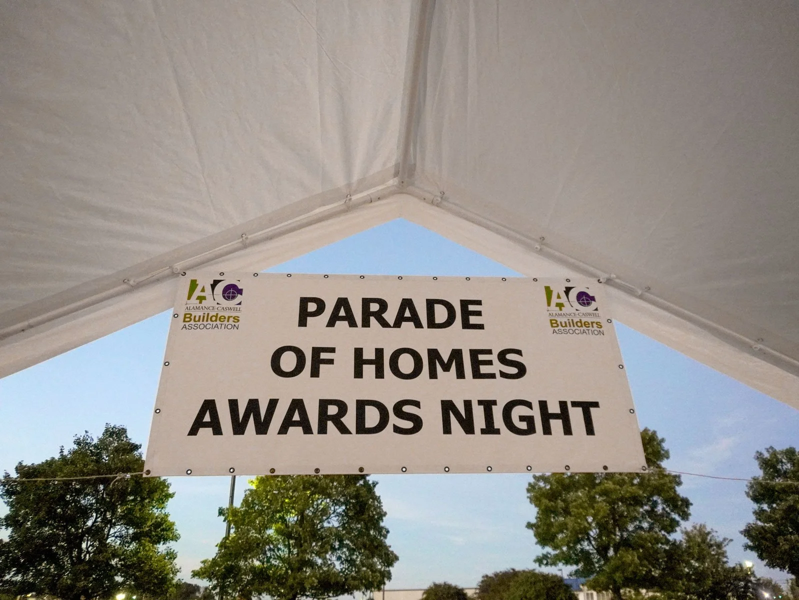 A banner hanging under a tent that reads 'Parade of Homes Awards Night' with the Alamance Caswell Builders Association logo on either side of the text. Trees and a blue sky are visible outside the tent.