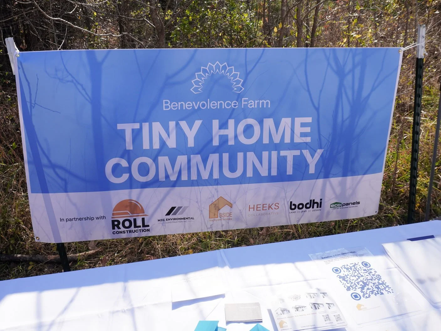 A printed signboard for Benevolence Farm Tiny Home Community outdoors with trees in the background. The sign features the farm's logo and names of partner organizations including Roll Construction, MEC Environmental, BSIDE, Heeks Collaborative, Bodhi, and Eco Panels.