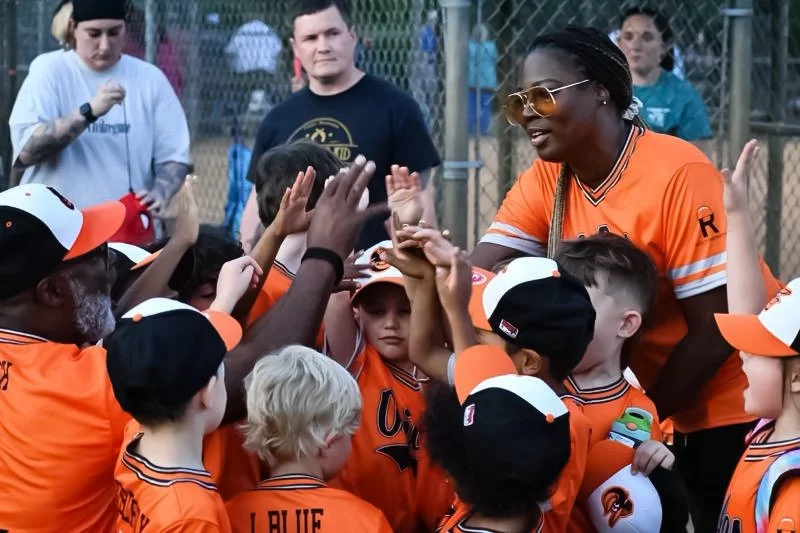 Youth baseball team in orange jerseys gather around coach for a high-five at the field during daytime.