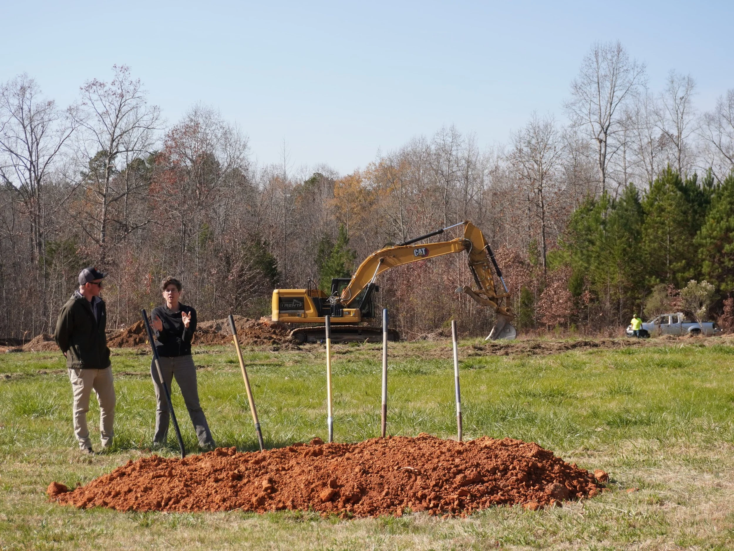 a man and woman talking in a field with an excavator in the background and four shovels in a pile of dirt in the foreground