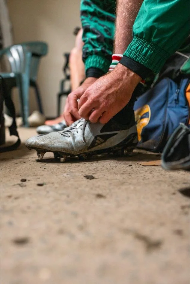 A person wearing green sportswear is tying the laces of a silver and black cleat on an indoor dirt or concrete floor, with other shoes and chairs in the background.
